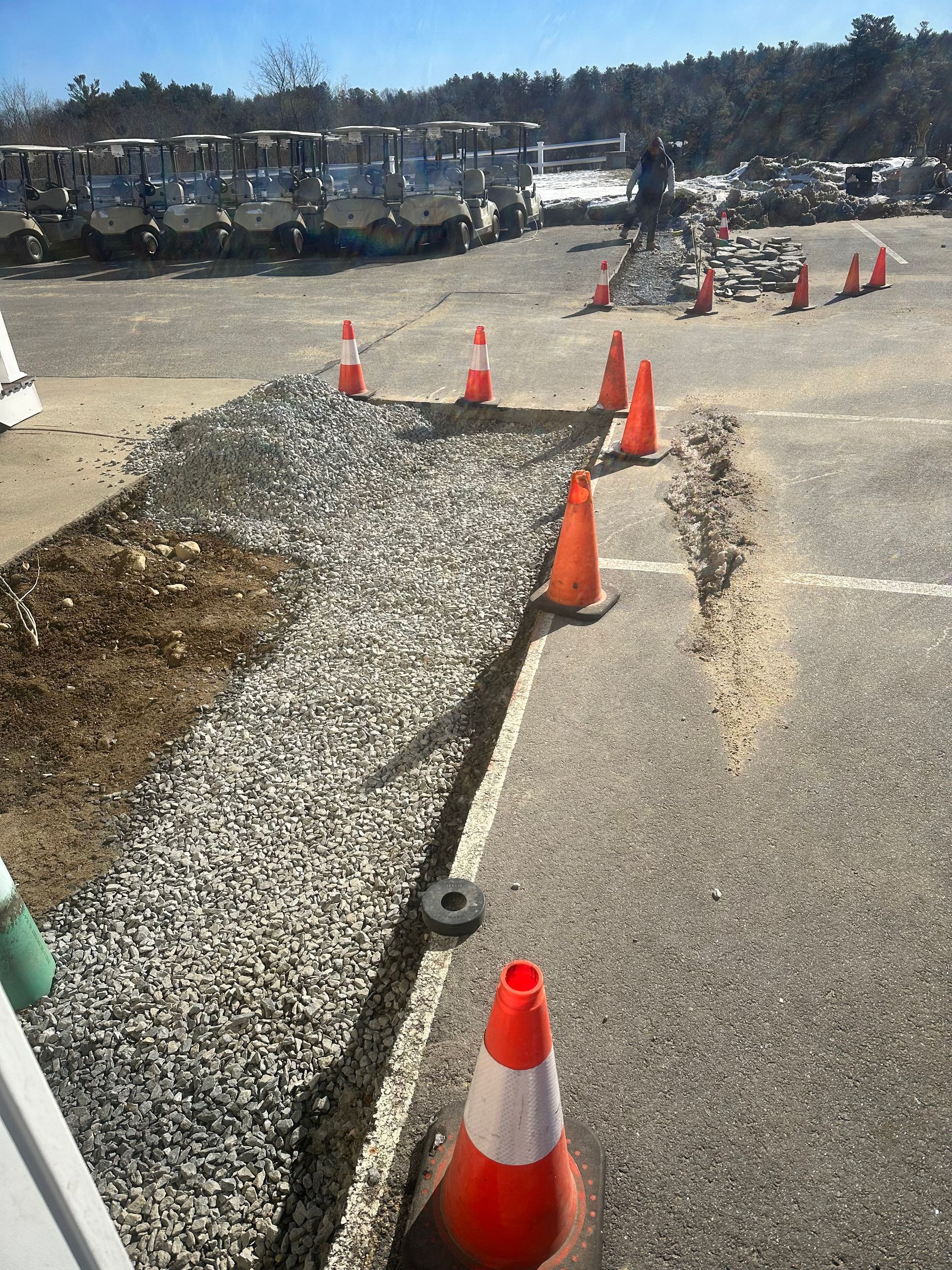 Orange traffic cones marking a gravel trench beside a paved lot, with construction equipment in the background