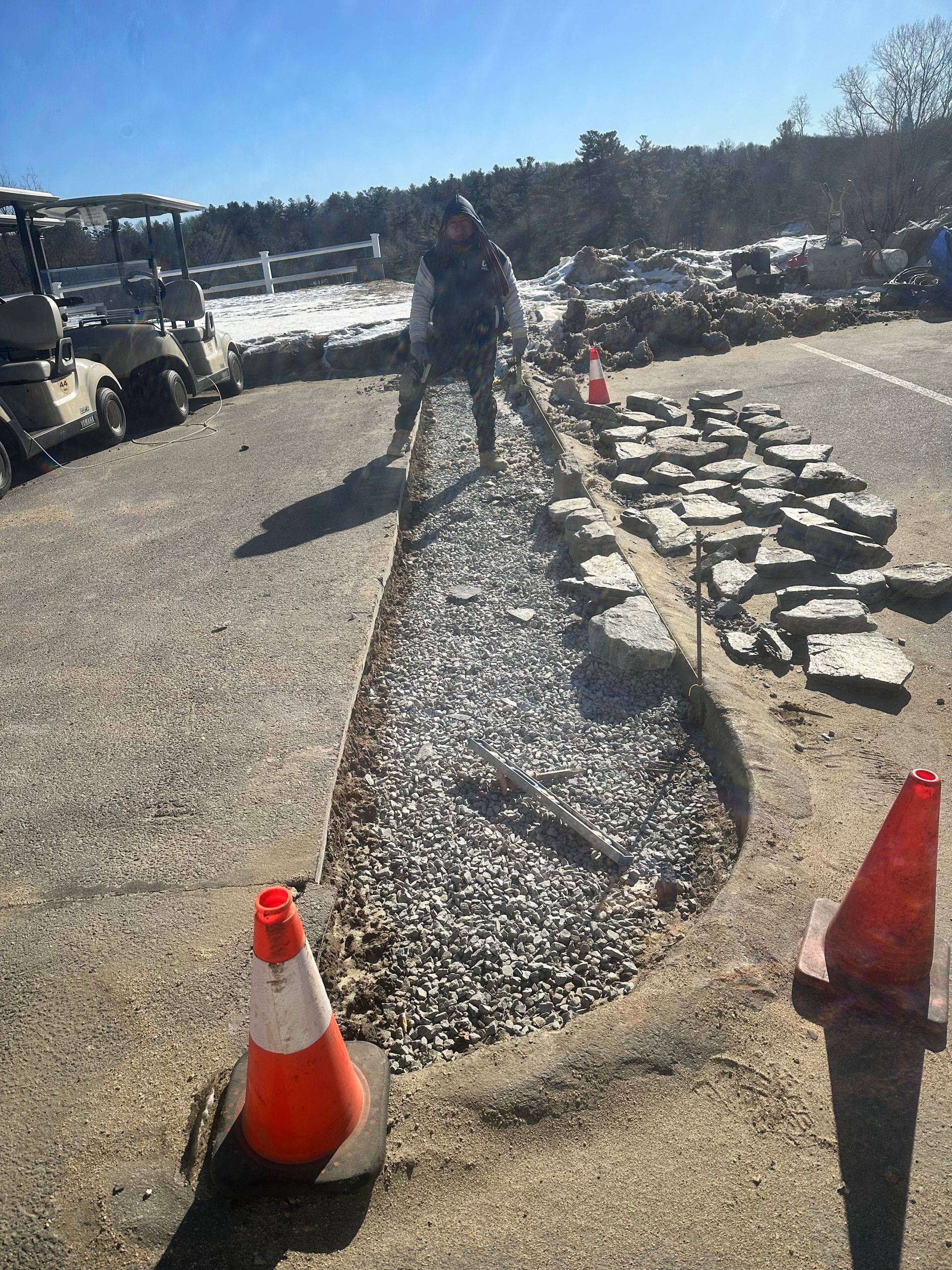 Roadwork scene with gravel trench, traffic cones, worker, and construction vehicle on a sunny roadside