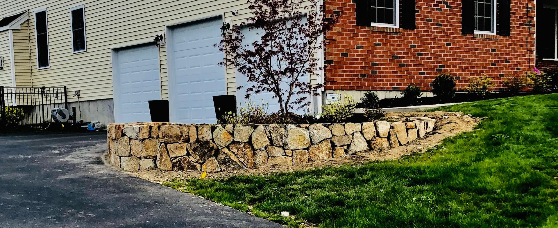 Stone retaining wall beside white garage doors and a brick house with a green lawn and driveway
