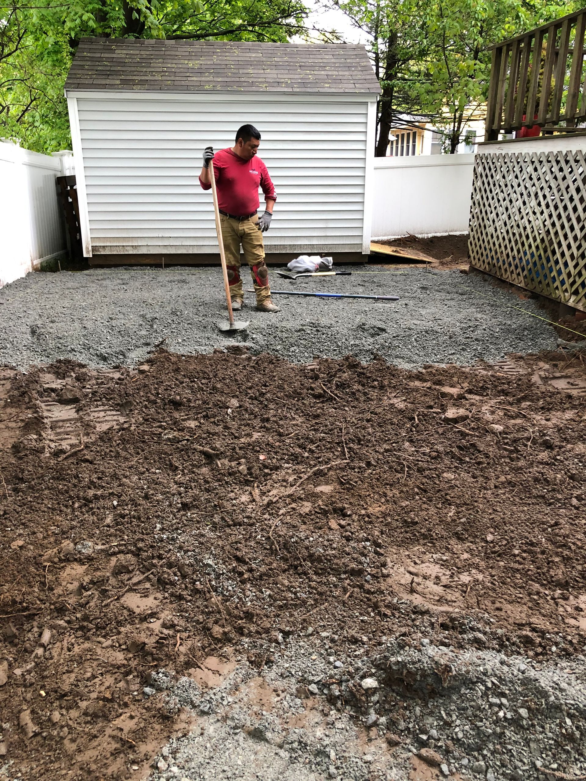 Person in a red shirt standing in a gravel yard in front of a white shed and trees