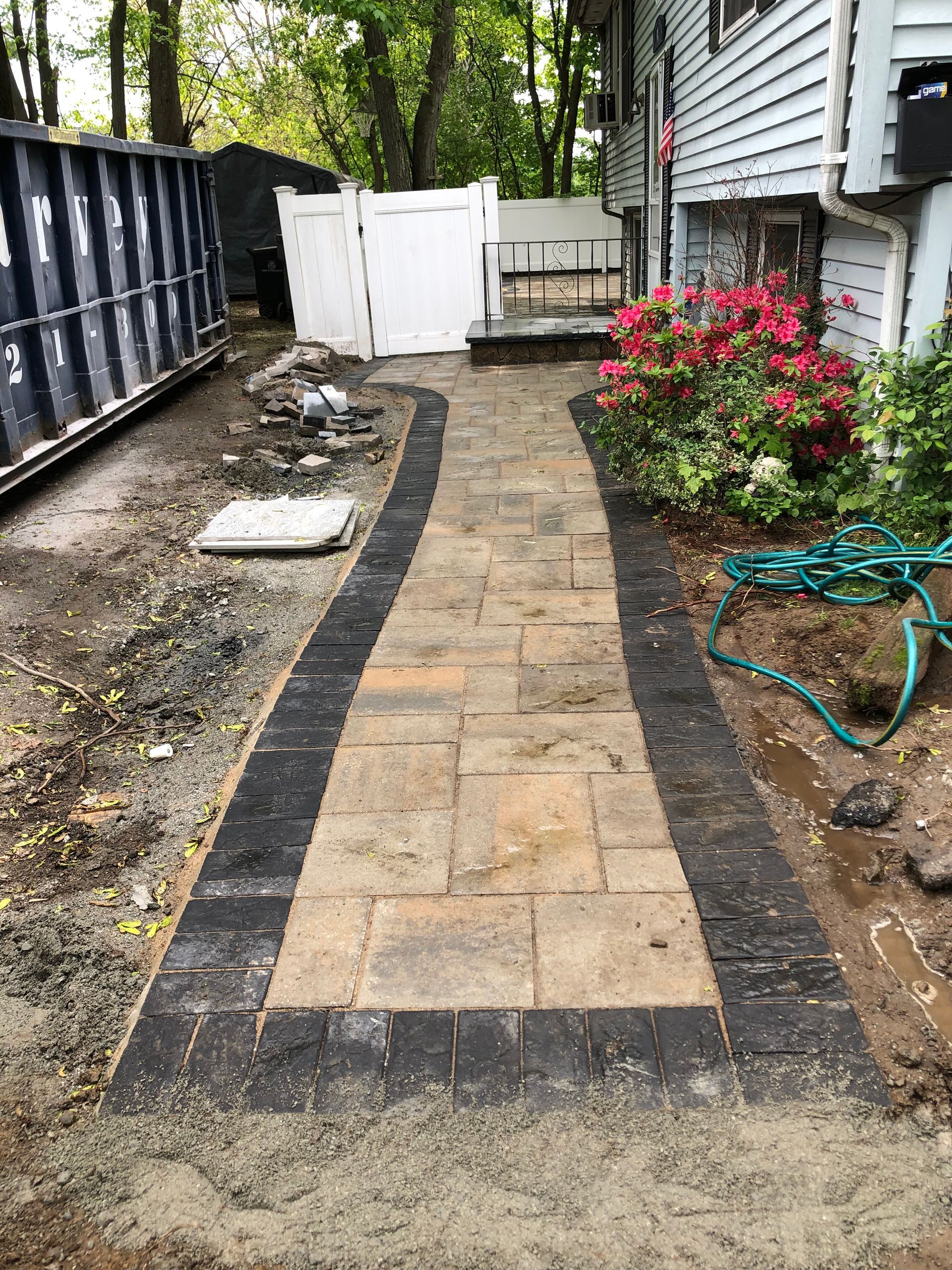 New brick walkway under construction beside a house, with dirt edges, fence, and garden hose.