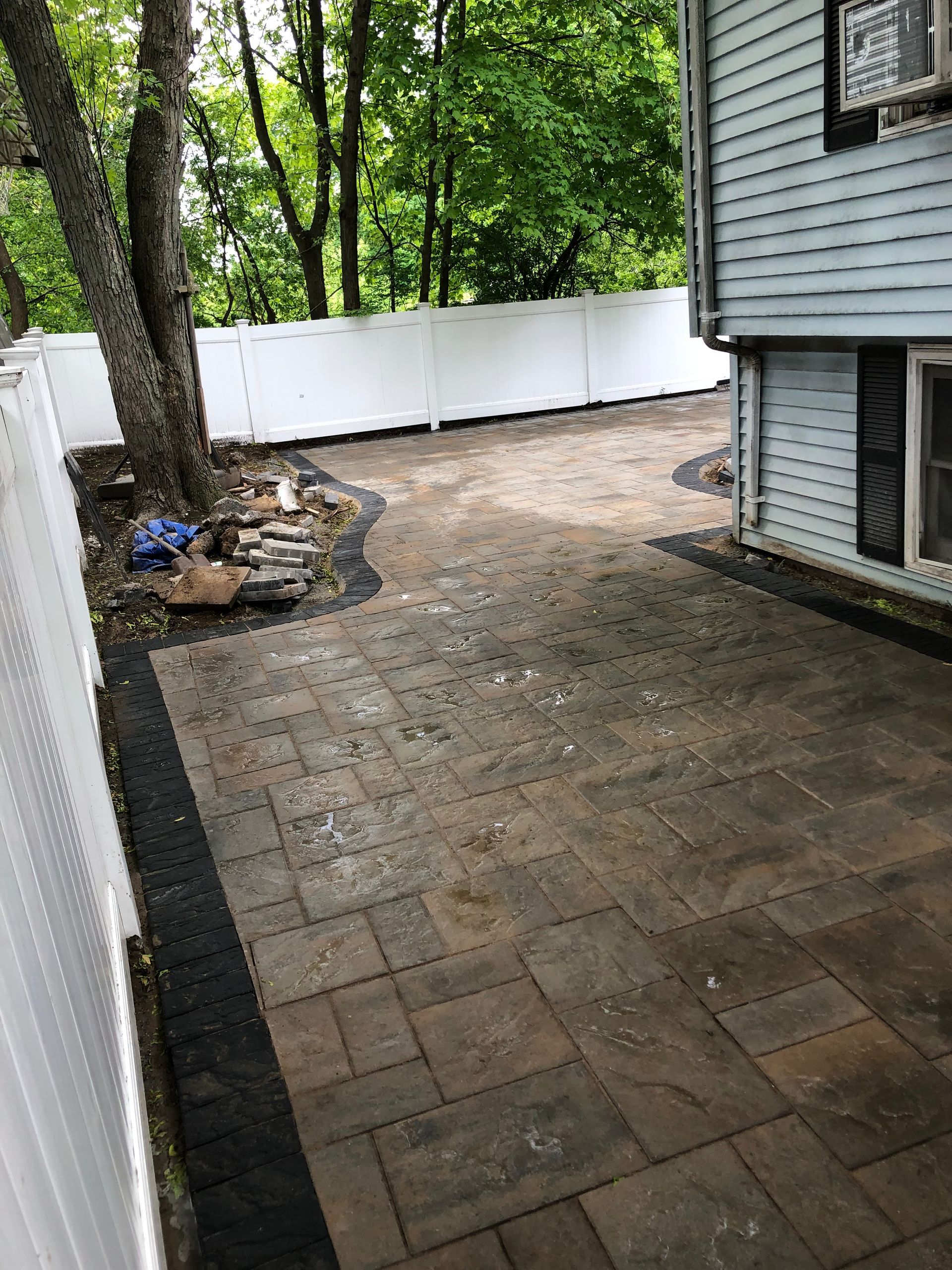 Backyard patio with stone pavers, white fence, trees, and the side of a house