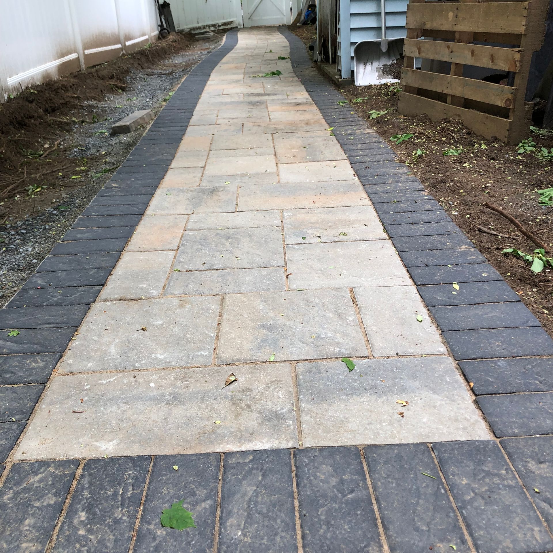 Narrow stone walkway with gray pavers between dirt borders beside a fence and building.