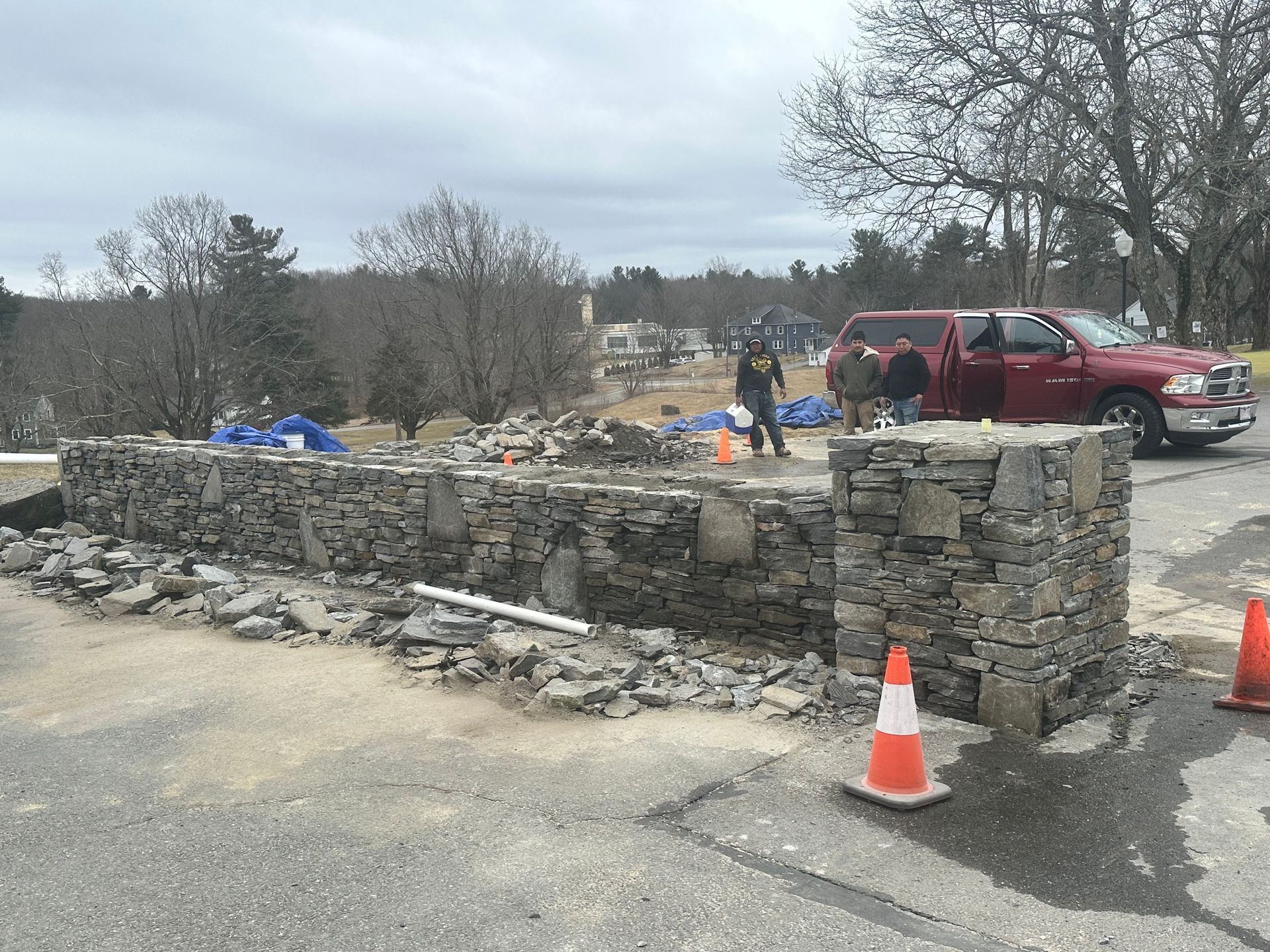 Stone roadside memorial wall with flowers and cones beside a red SUV in a parking area