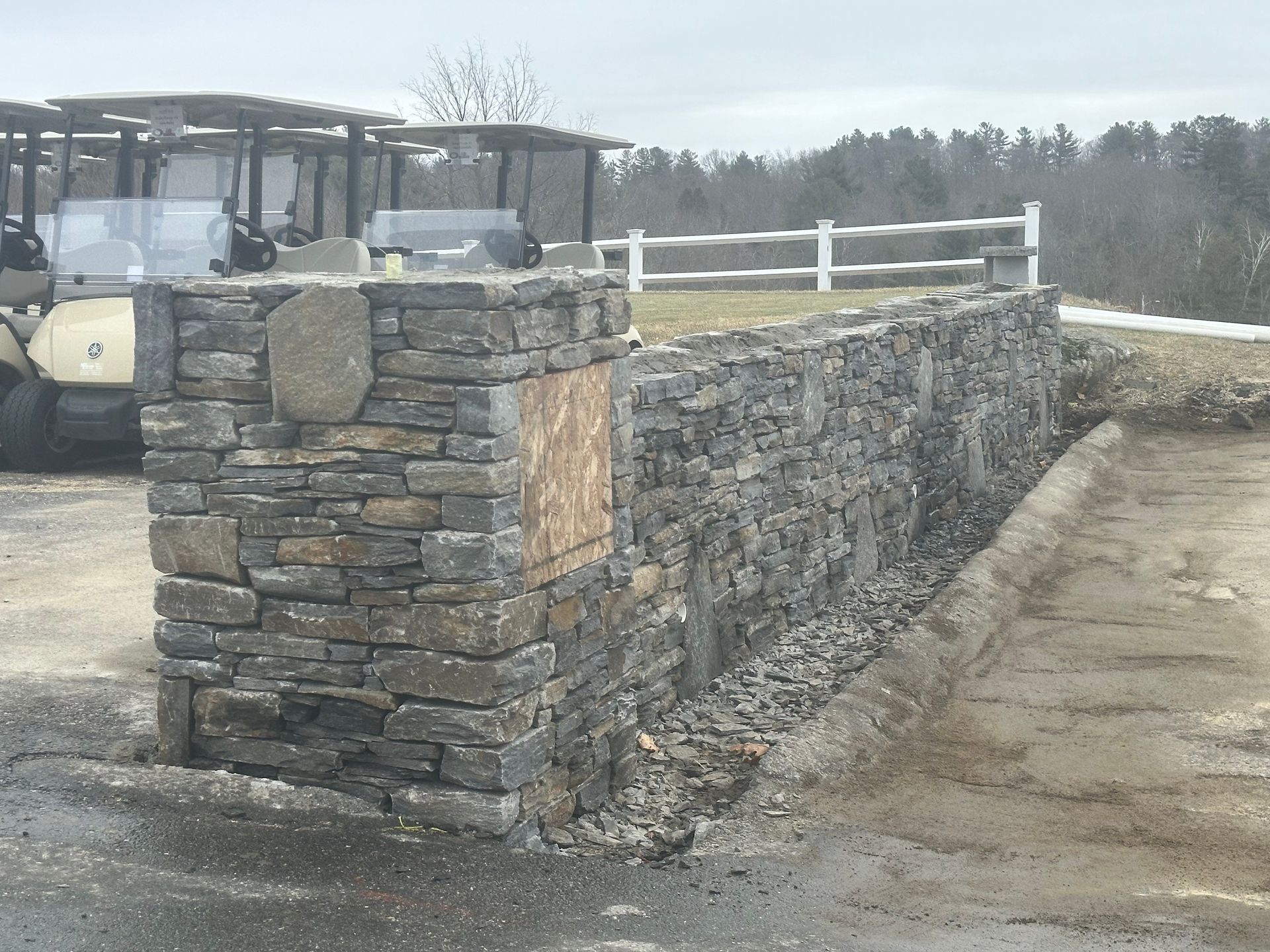 Stone retaining wall beside a gravel drive, with a forklift in the background under cloudy skies.