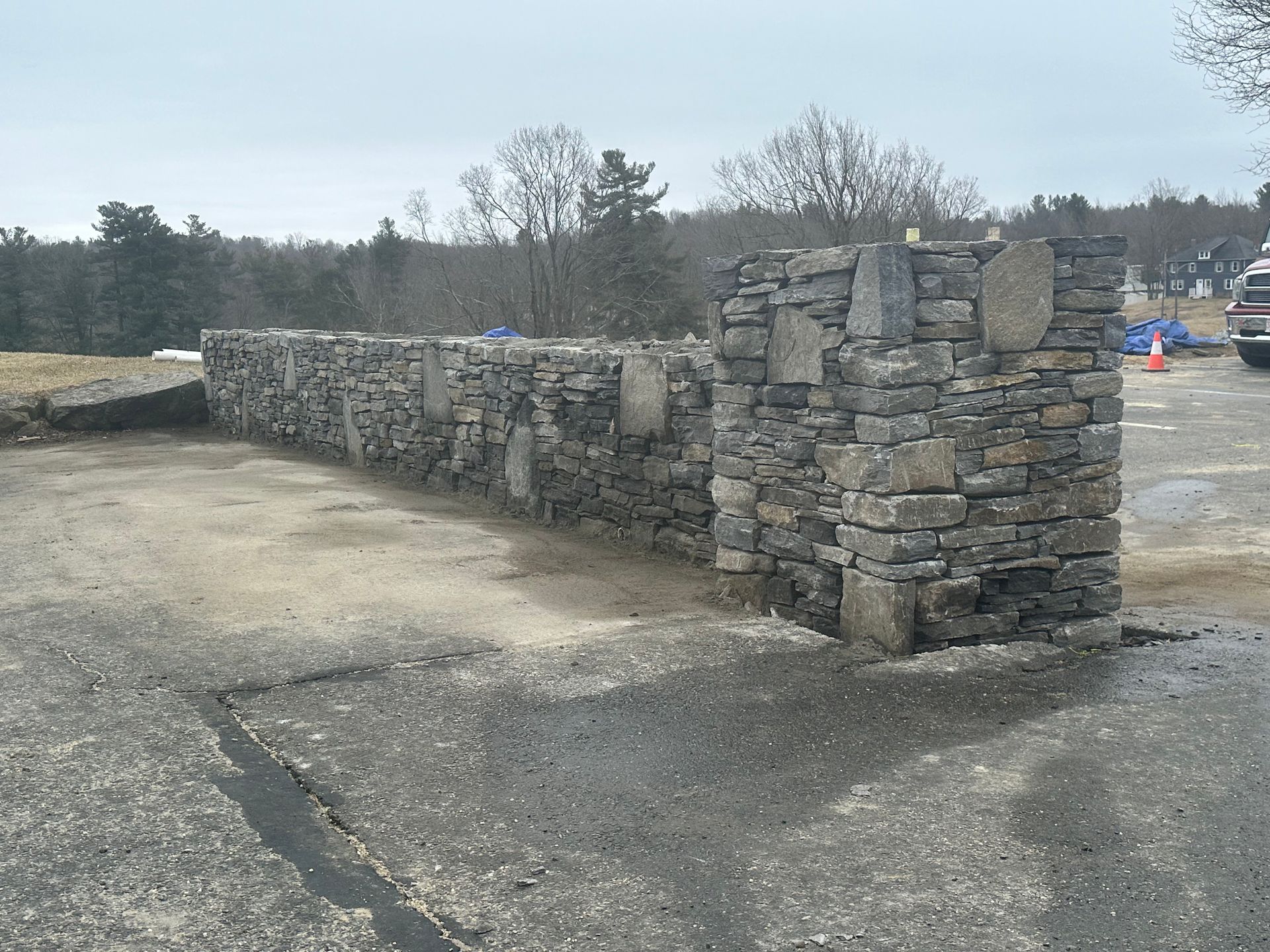 Stone wall ruins beside a dirt lot under an overcast sky, with trees in the background
