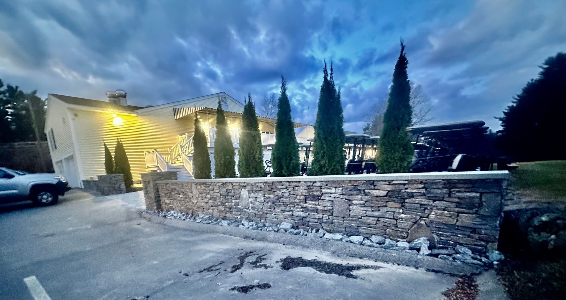 Stone wall and tall cypress trees outside a warmly lit building at dusk