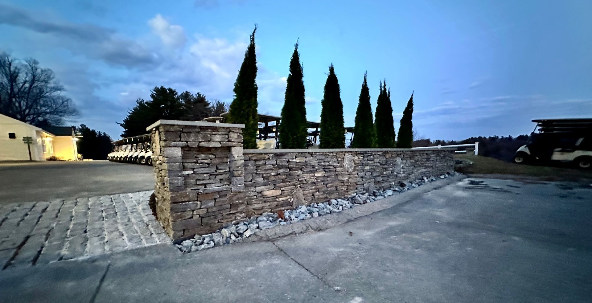 Stone retaining wall with tall cypress trees beside a paved drive at dusk
