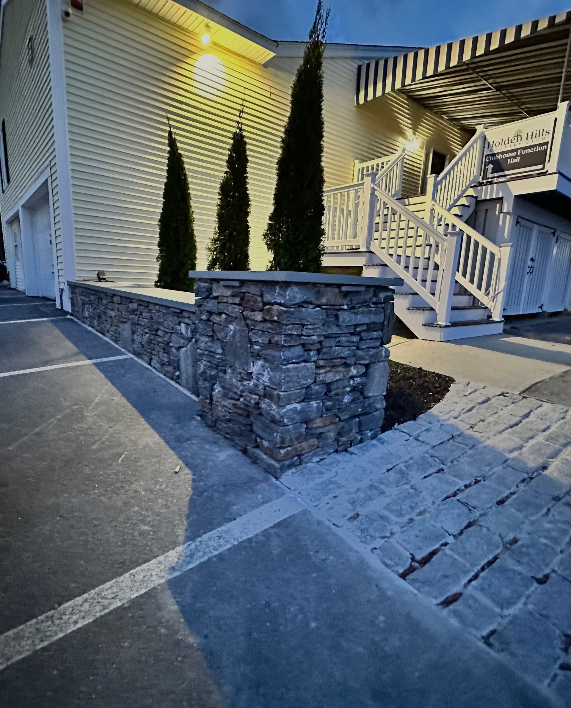 Stone planter wall with tall evergreens beside a house porch and stairs at dusk