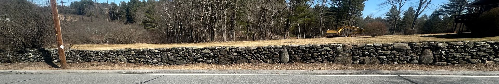 Snow-lined stone wall beside a road with leafless trees and dry grass under a clear sky
