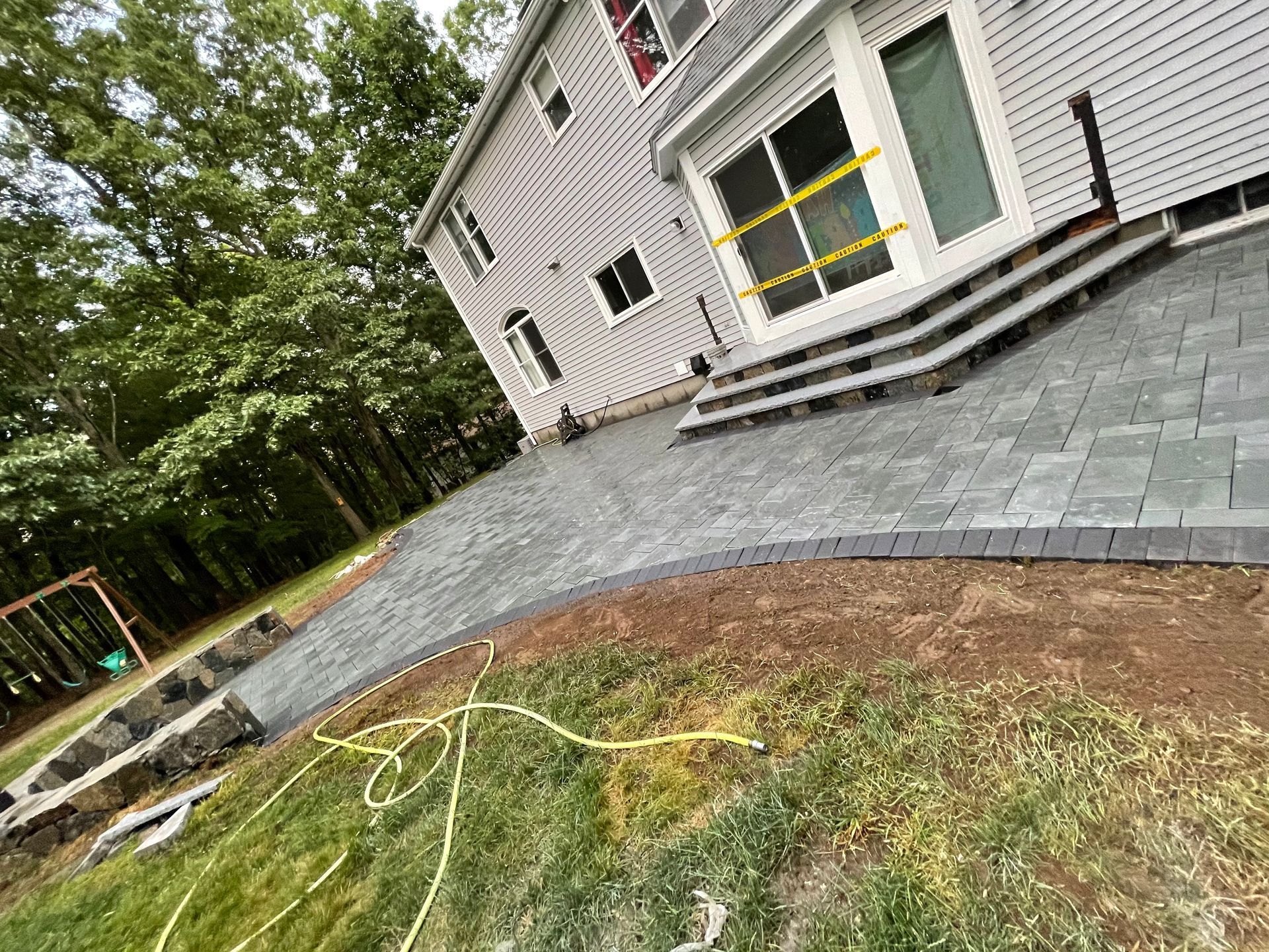 Tilted view of a house exterior with a stone patio, grassy yard, and trees nearby