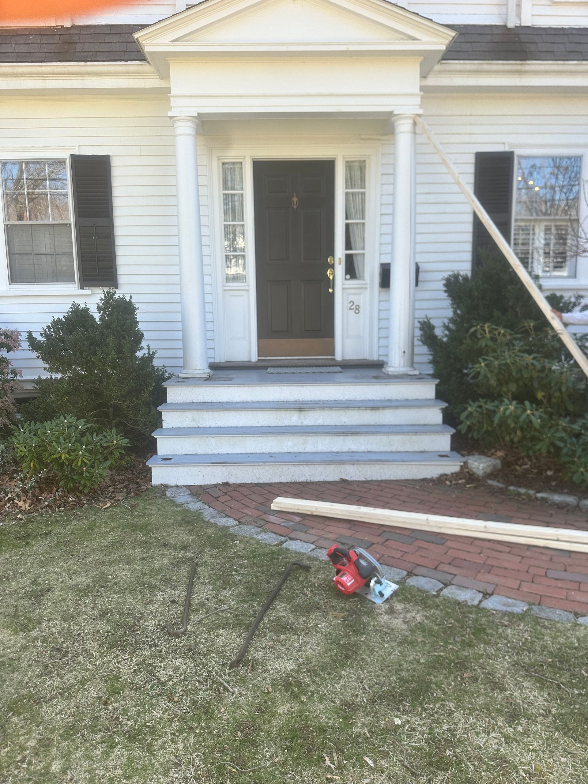 Front entrance of a white house with steps, brick walkway, shrubs, and a red leaf blower on the lawn