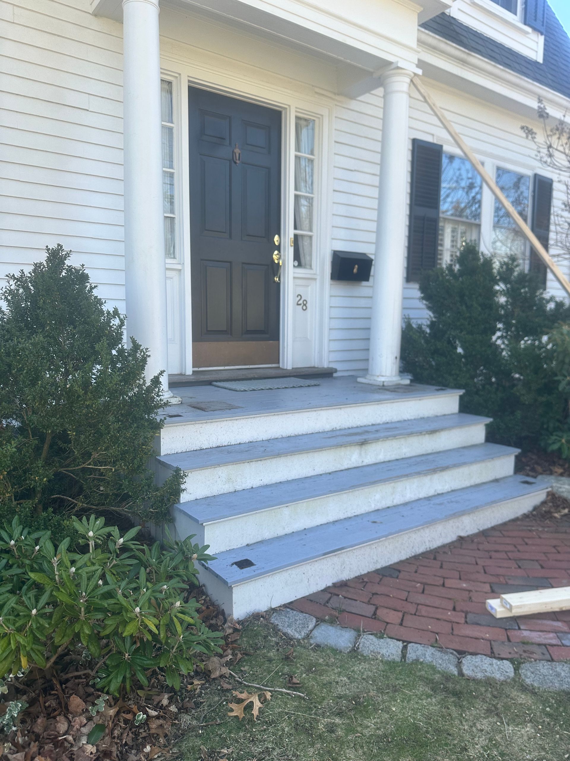 Front porch with white steps, dark door, and shrubs beside a brick walkway