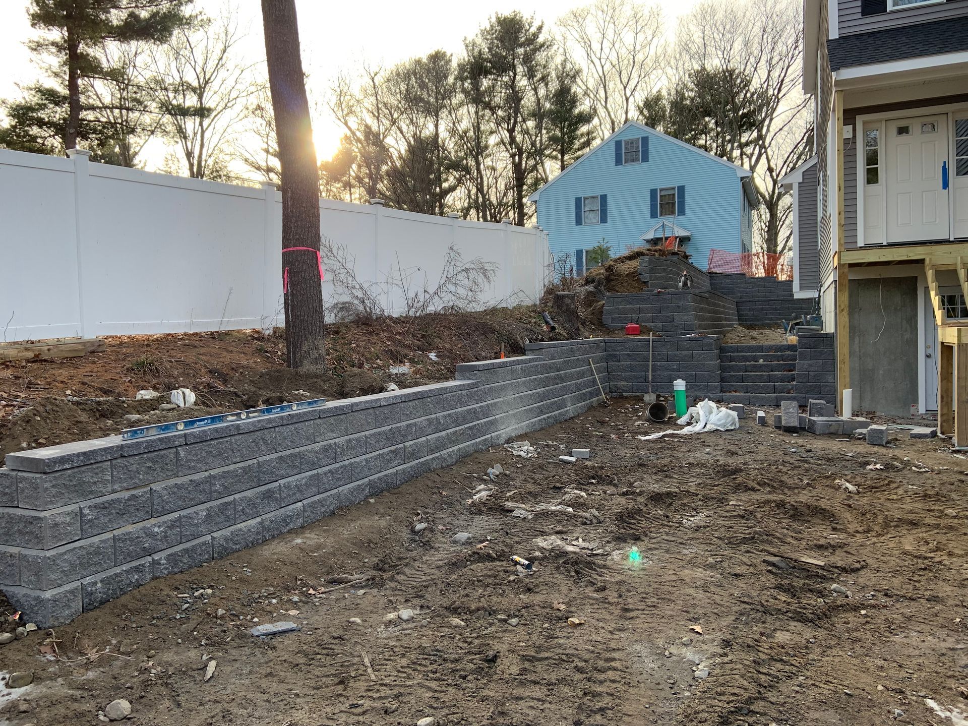 Backyard retaining wall under construction beside a house, with dirt, steps, and a blue shed in the background