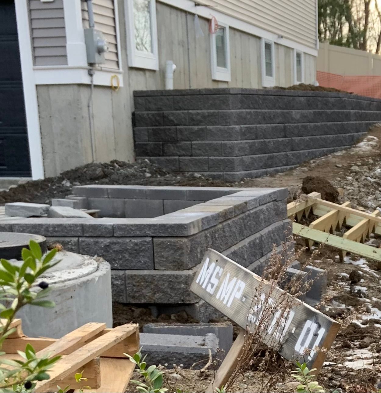 Backyard construction site beside a house, with stacked retaining wall blocks and muddy debris.