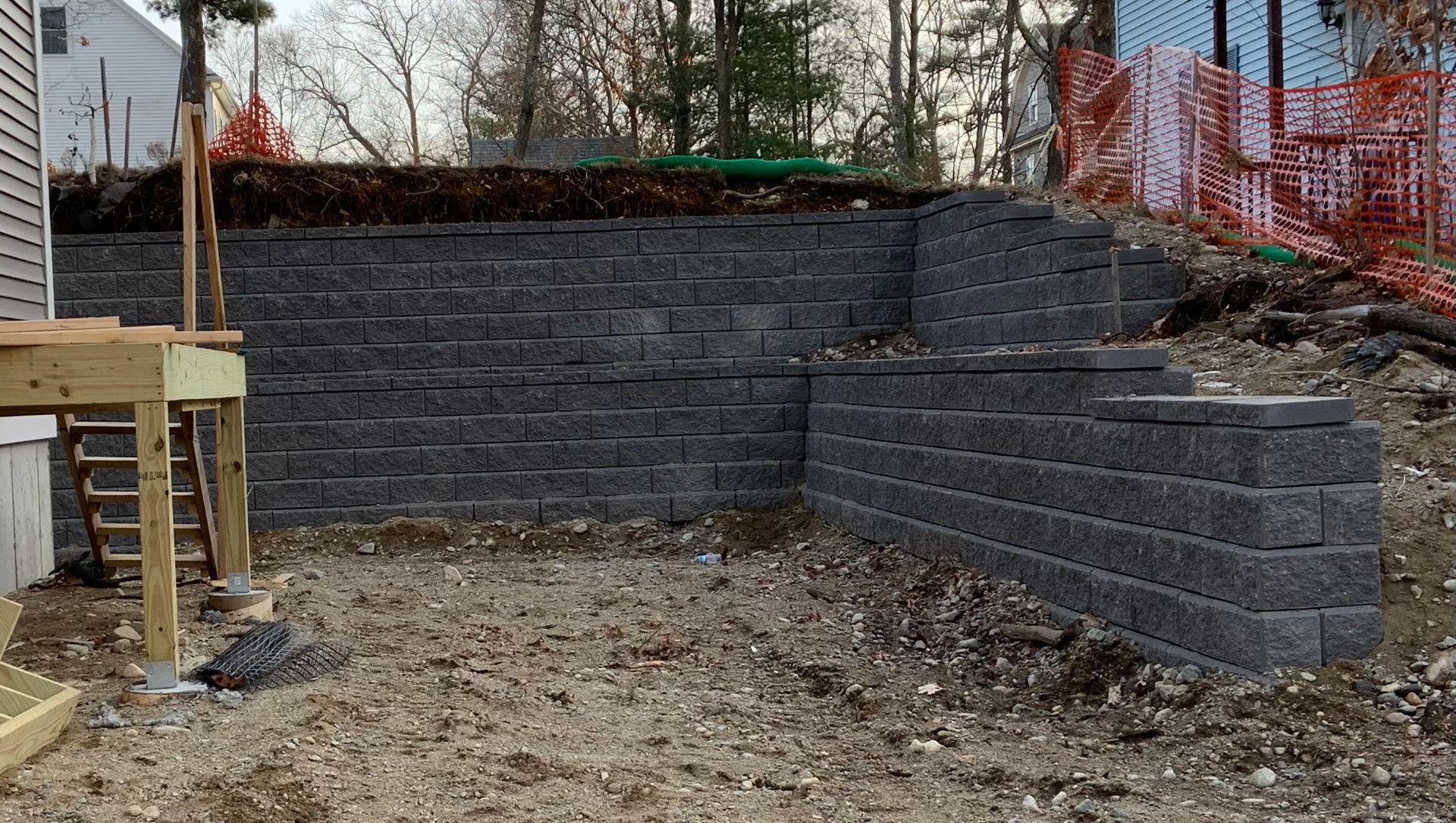 Retaining wall made of gray blocks under construction in a dirt yard, with orange safety fencing nearby