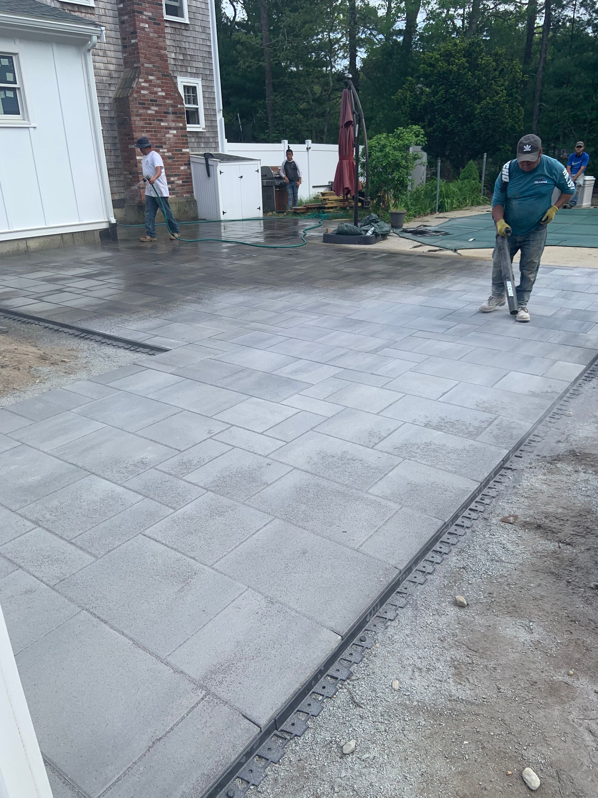 Wet concrete patio beside a brick house, with two workers on a rainy day.