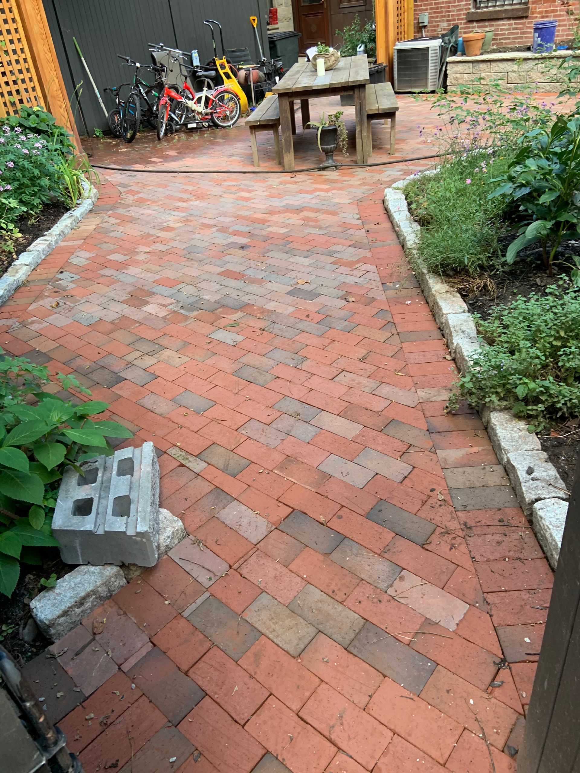 Brick-paved garden path leading to a patio table, bordered by plants and tools in the background