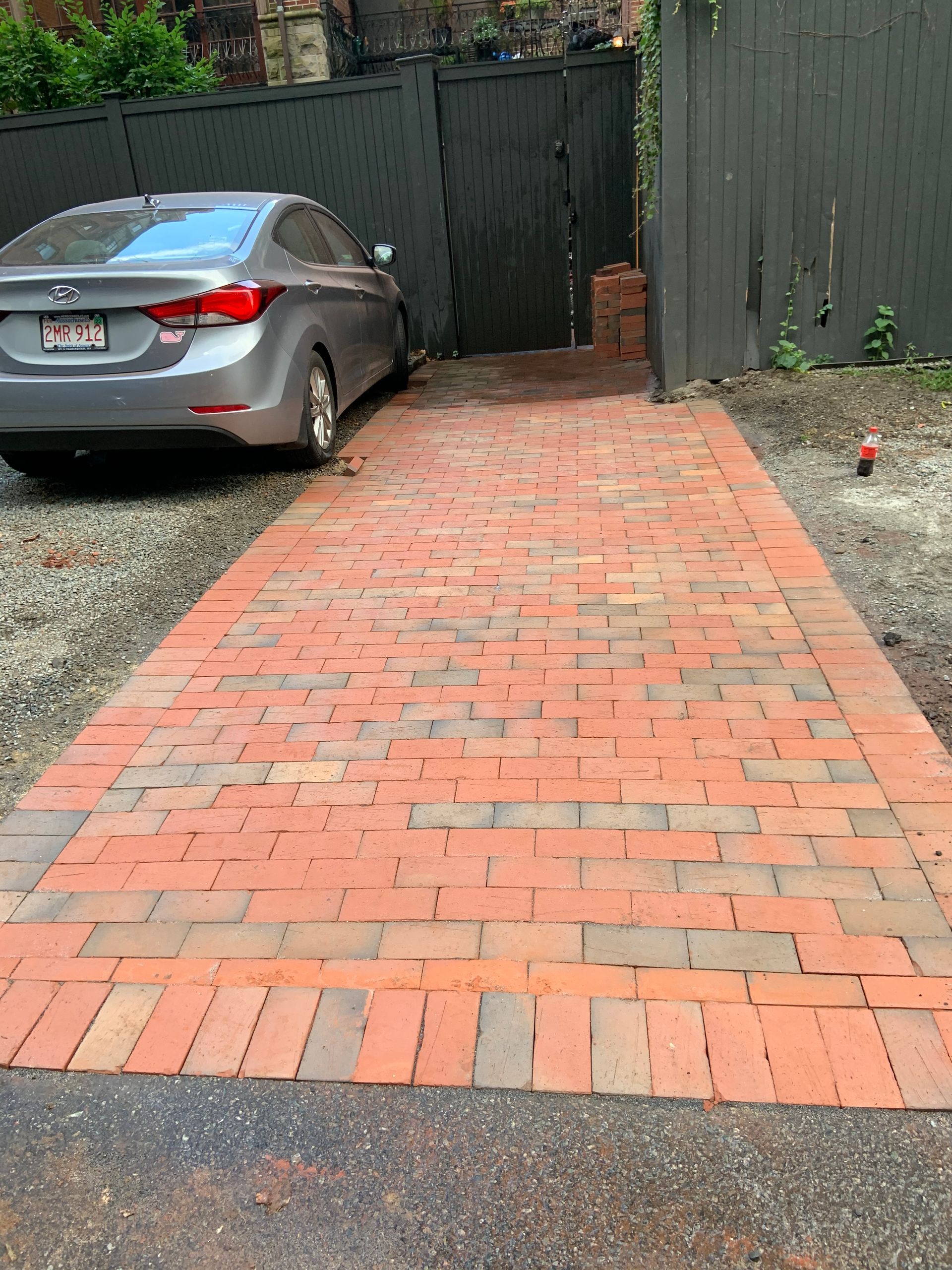 Red brick driveway leading to a dark green gate, with a gray car parked on the left.
