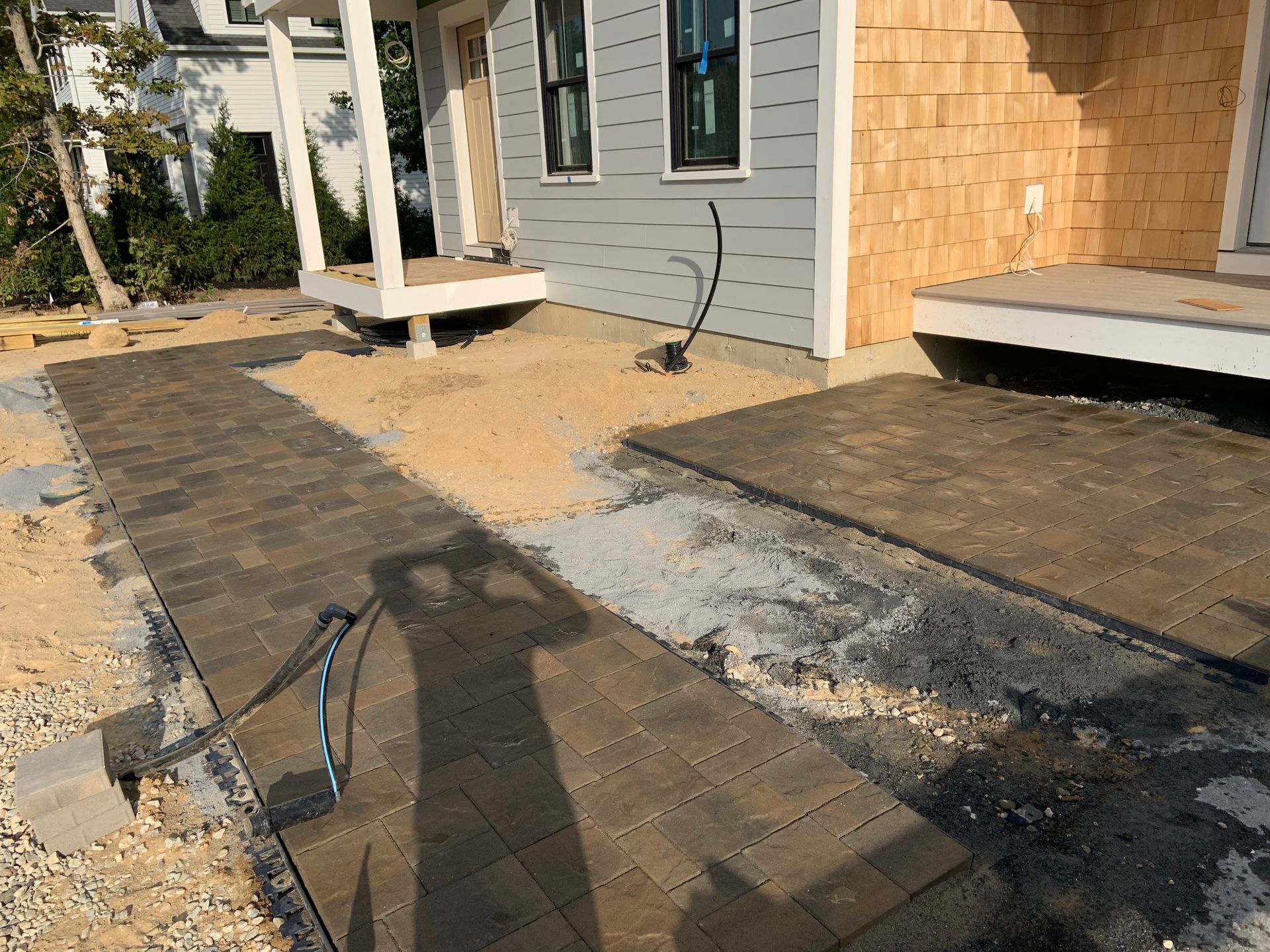 Partially paved walkway under construction beside a house, with dirt, gravel, and a porch deck nearby