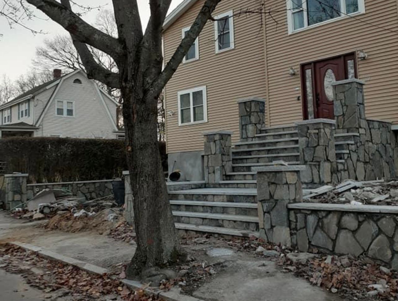 Brick house with stone porch wall, front steps, tree, and sidewalk in a residential neighborhood