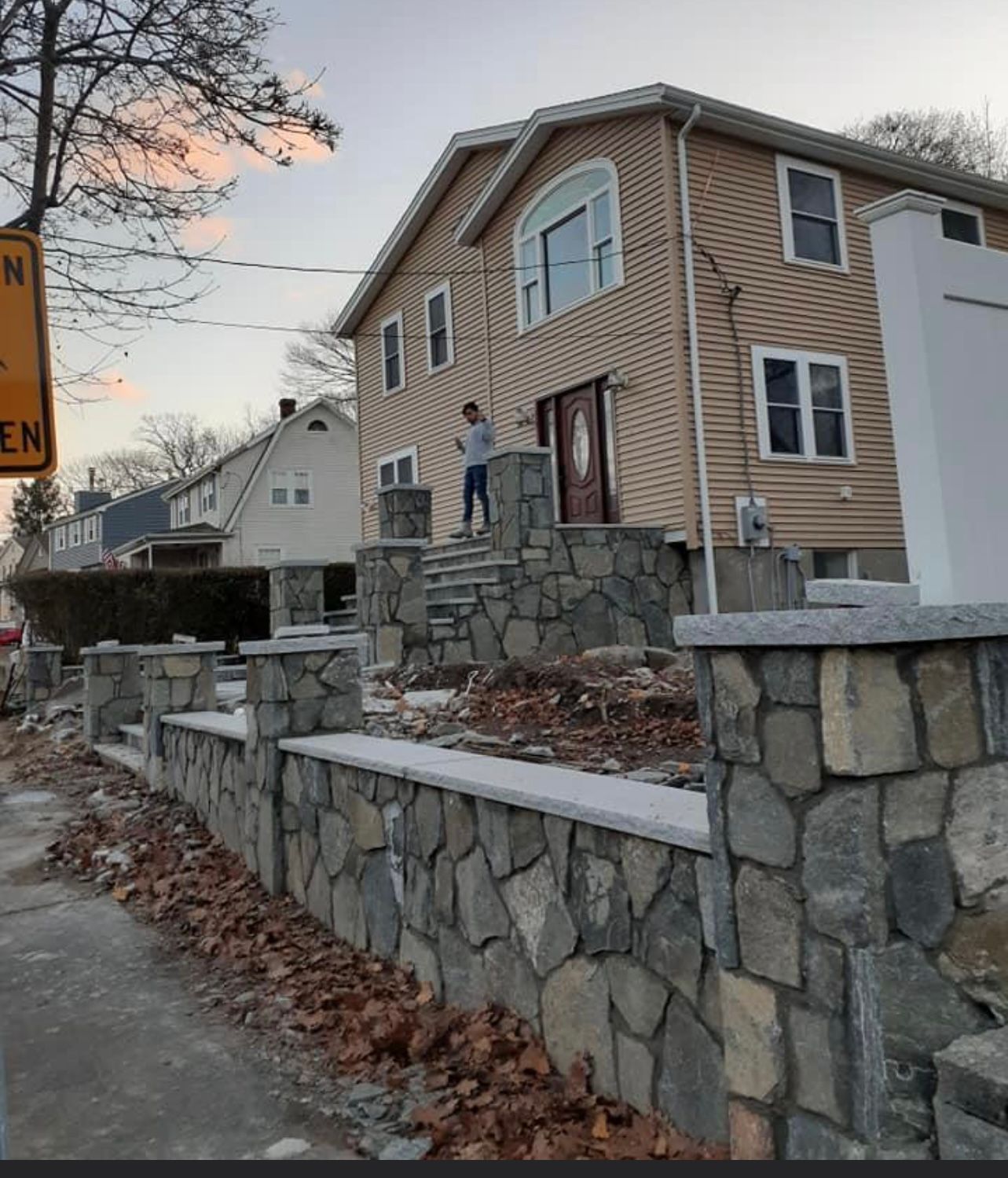 Stone retaining wall and beige house on a sloped street, with a person standing on the front steps.