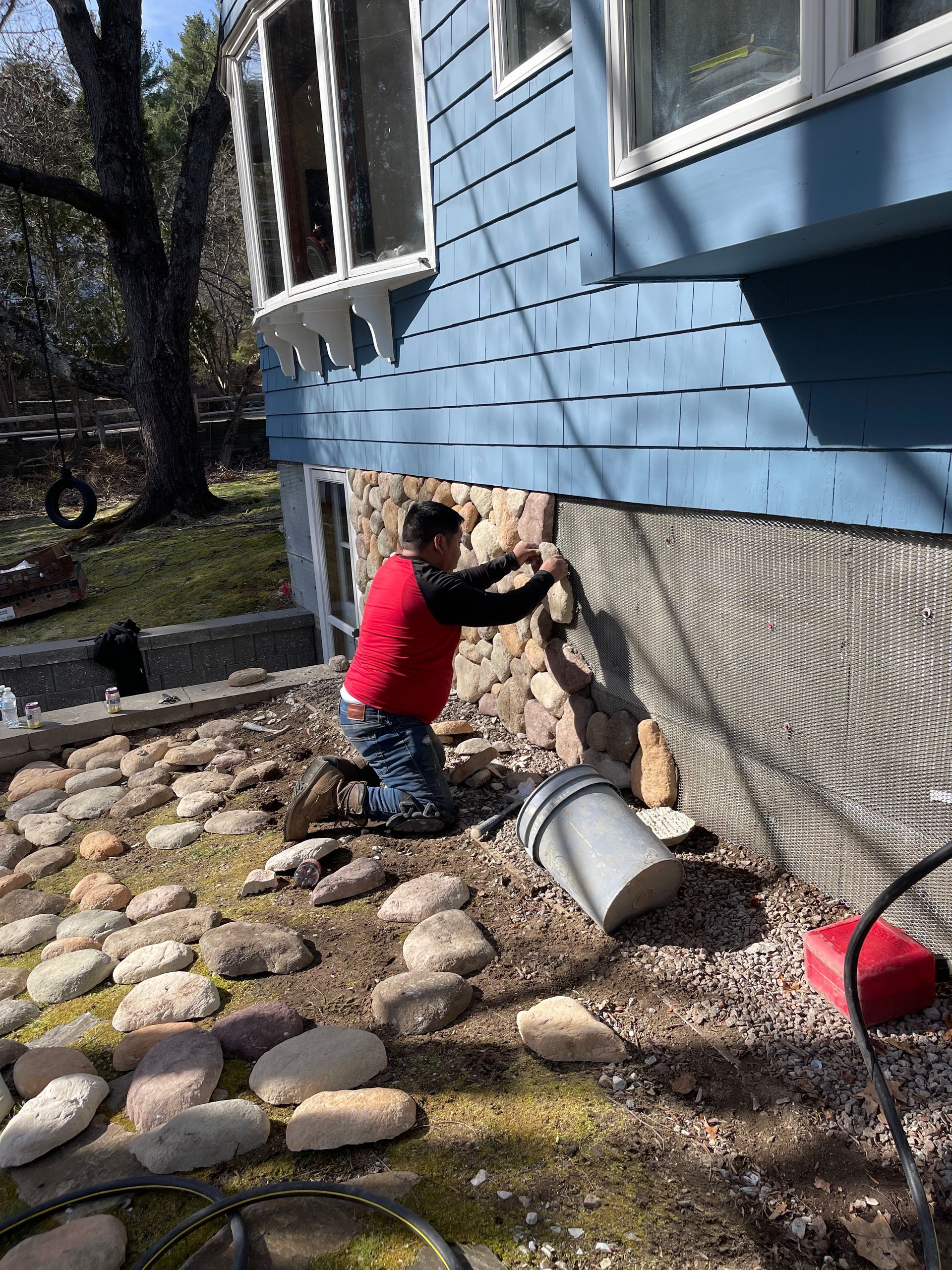 Person in red shirt working on the foundation of a blue house beside a rock garden.