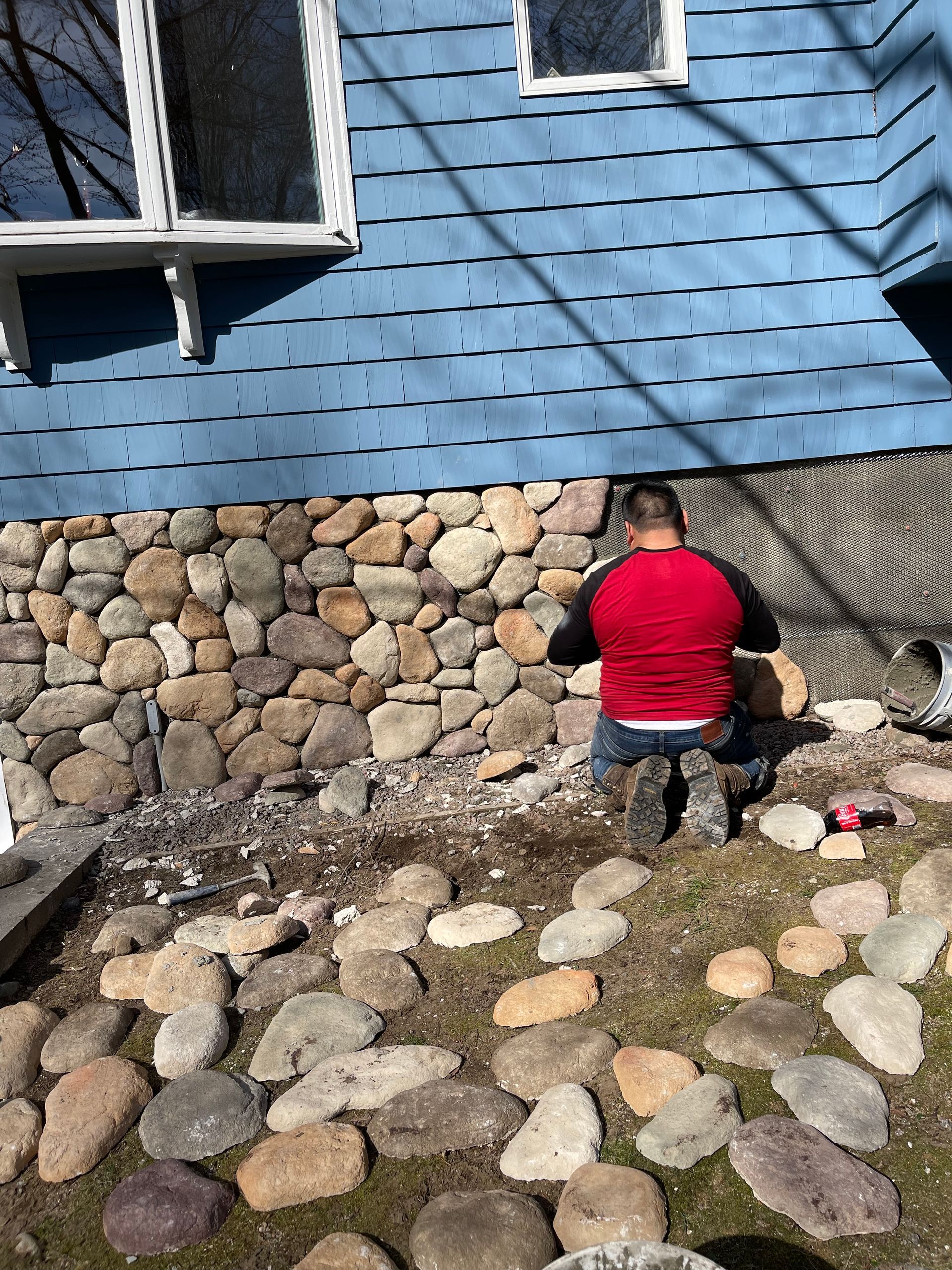 Person kneeling beside a blue house, working on a stone and mesh foundation wall.