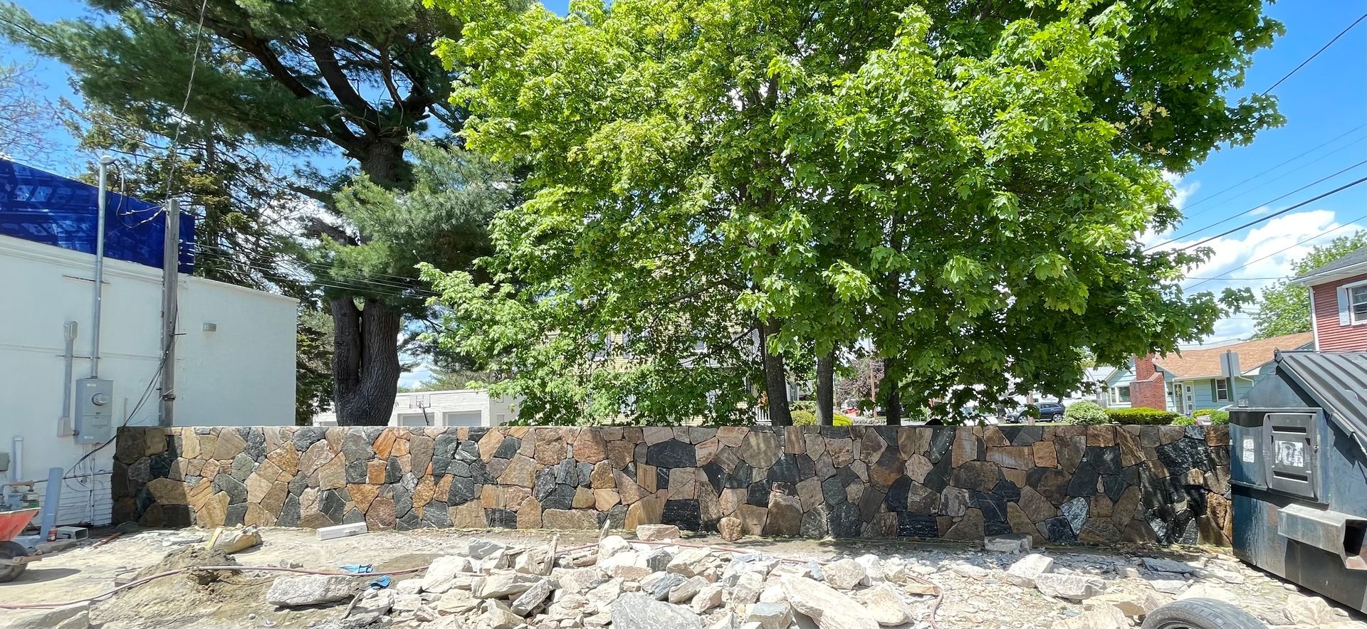 Stone wall and rubble in a sunny outdoor area with trees, a blue building, and a parked truck