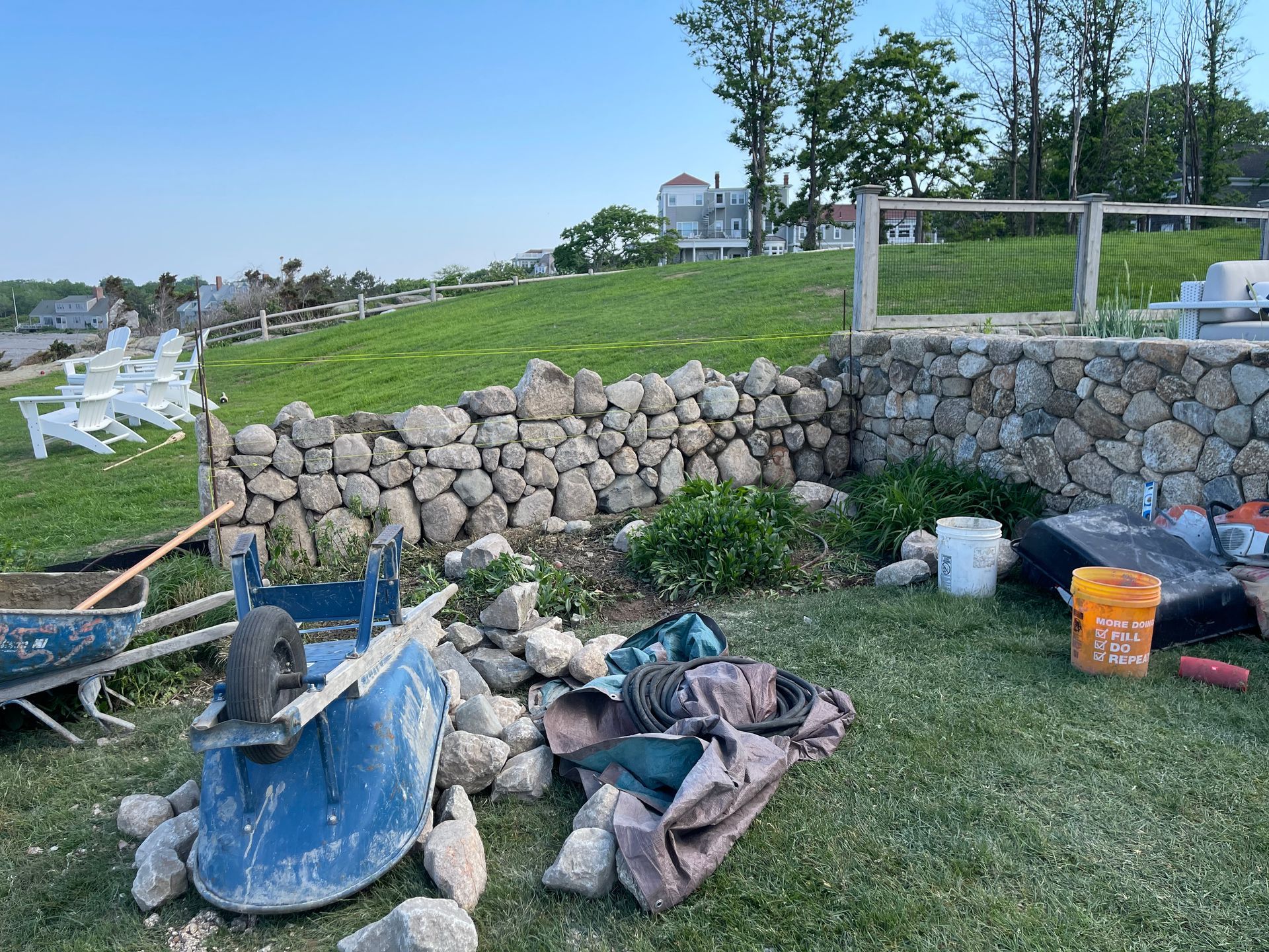 Garden work area with wheelbarrow, tools, rocks, and stone wall on grassy slope under a cloudy sky