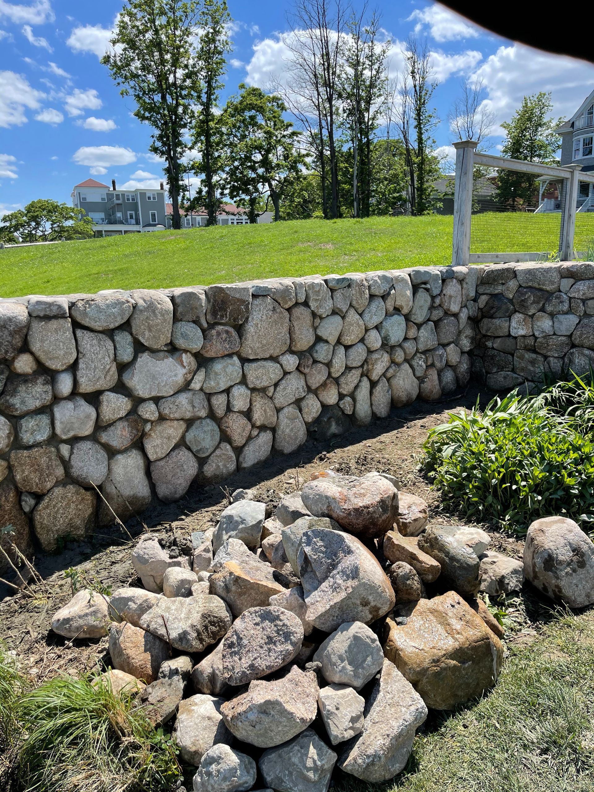 Stone retaining wall with stacked rocks, green lawn, and trees under a blue sky