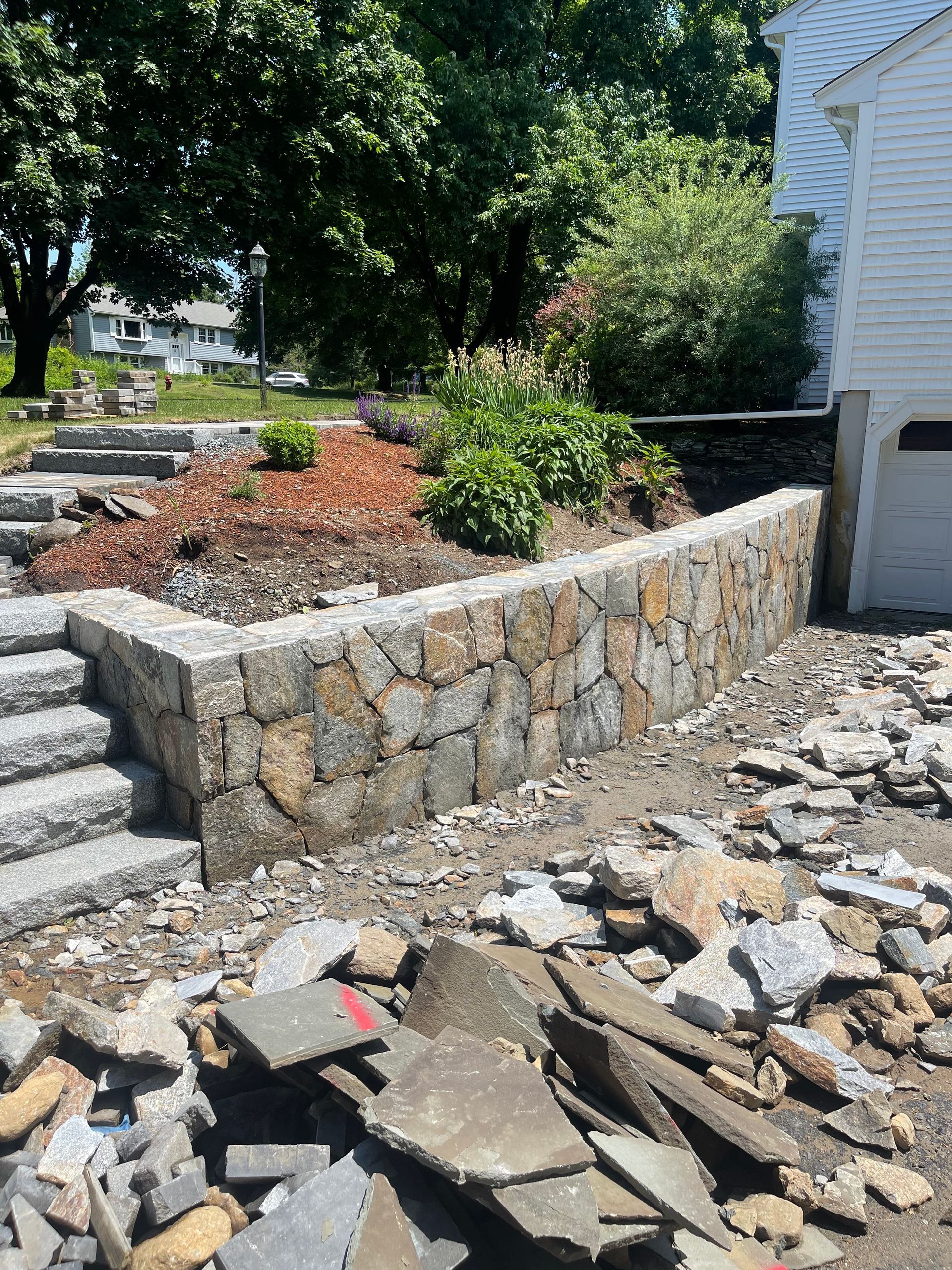 Stone retaining wall under construction beside house, with broken pavers and debris in the foreground