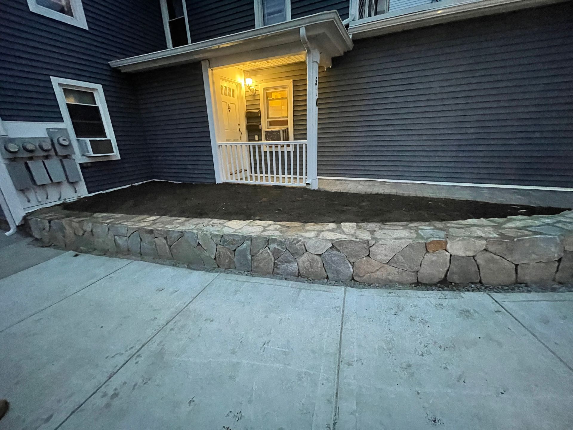 Stone retaining wall and small porch with lit doorway beside a concrete sidewalk at dusk