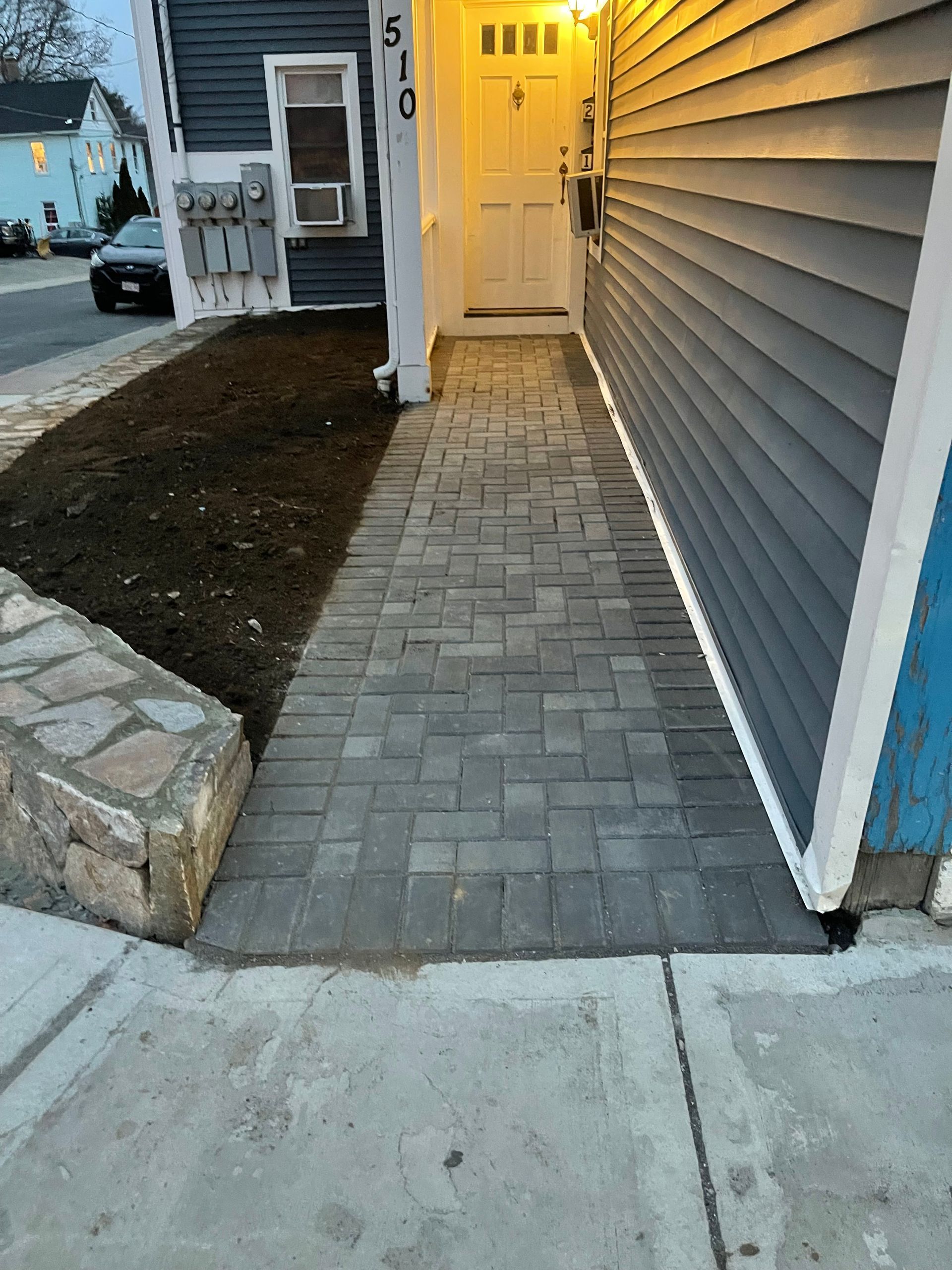 Brick walkway leading to a lit front door beside gray siding at dusk
