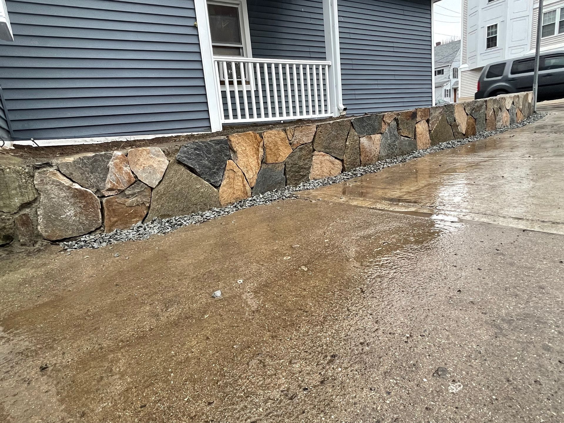 Wet concrete driveway beside a house with a stone retaining wall and white porch railing.