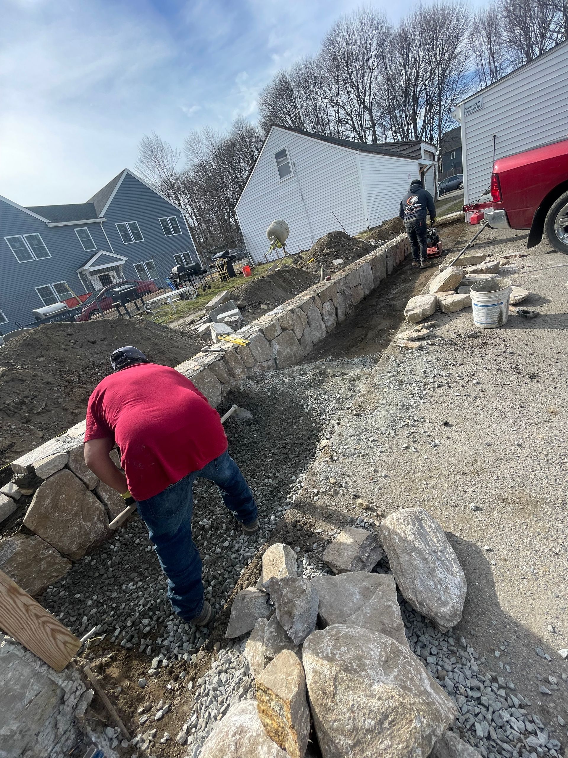 Workers in a gravel trench beside houses, with stone border wall and red truck nearby