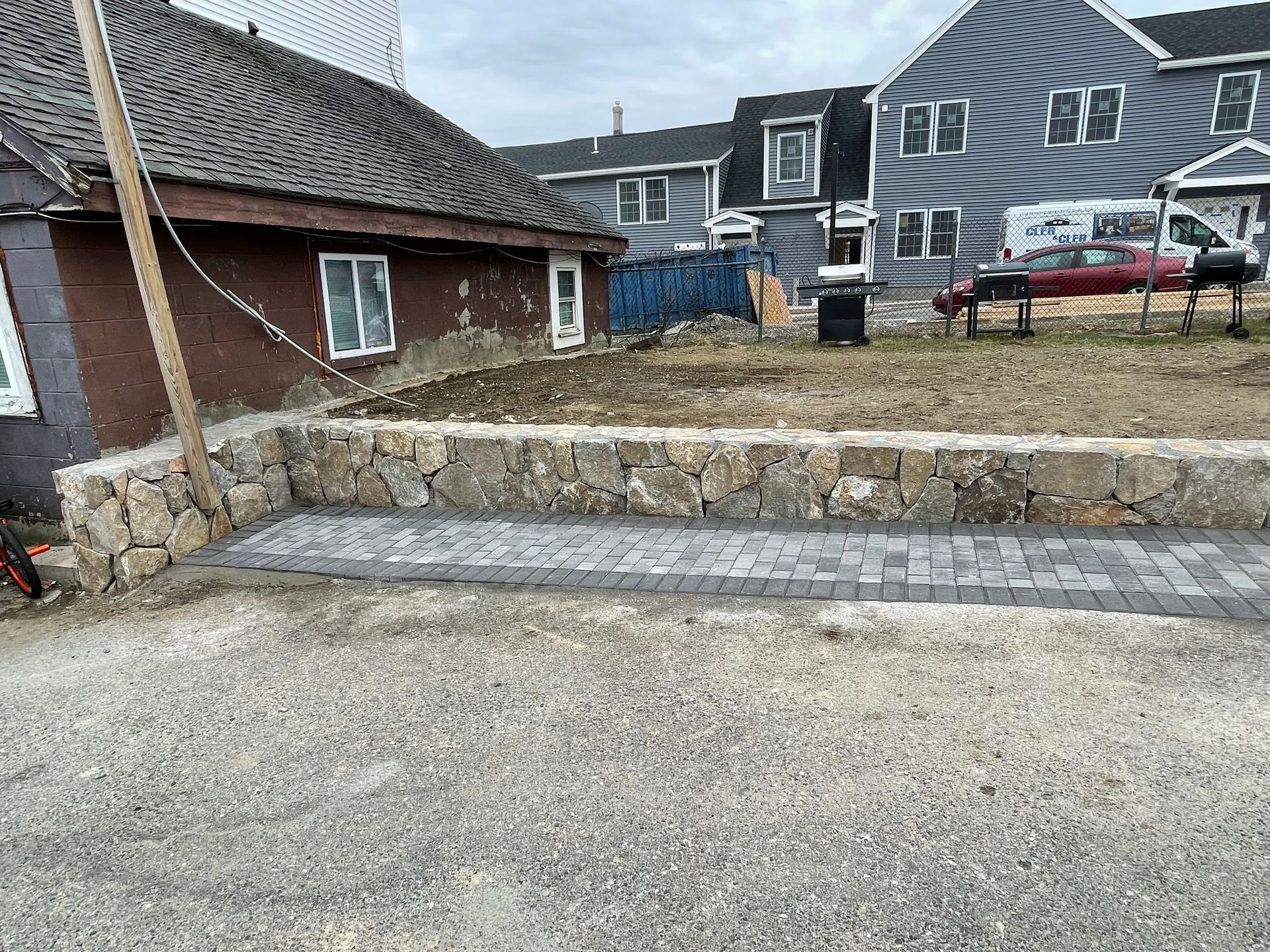 Stone retaining wall and steps beside a gravel lot, with houses and a parked car in the background