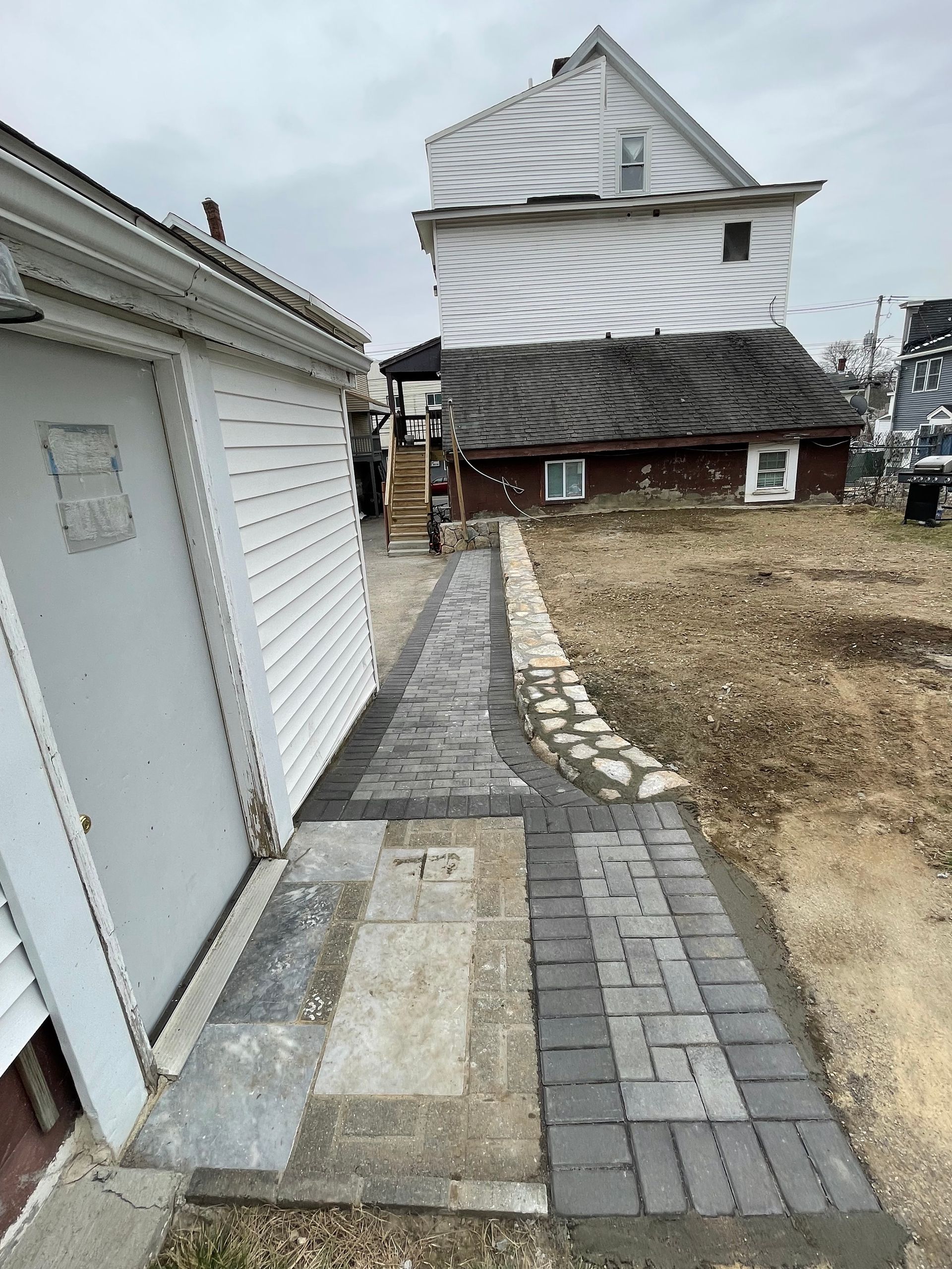 Narrow paved walkway between white buildings leading to a brown-roofed house in a yard