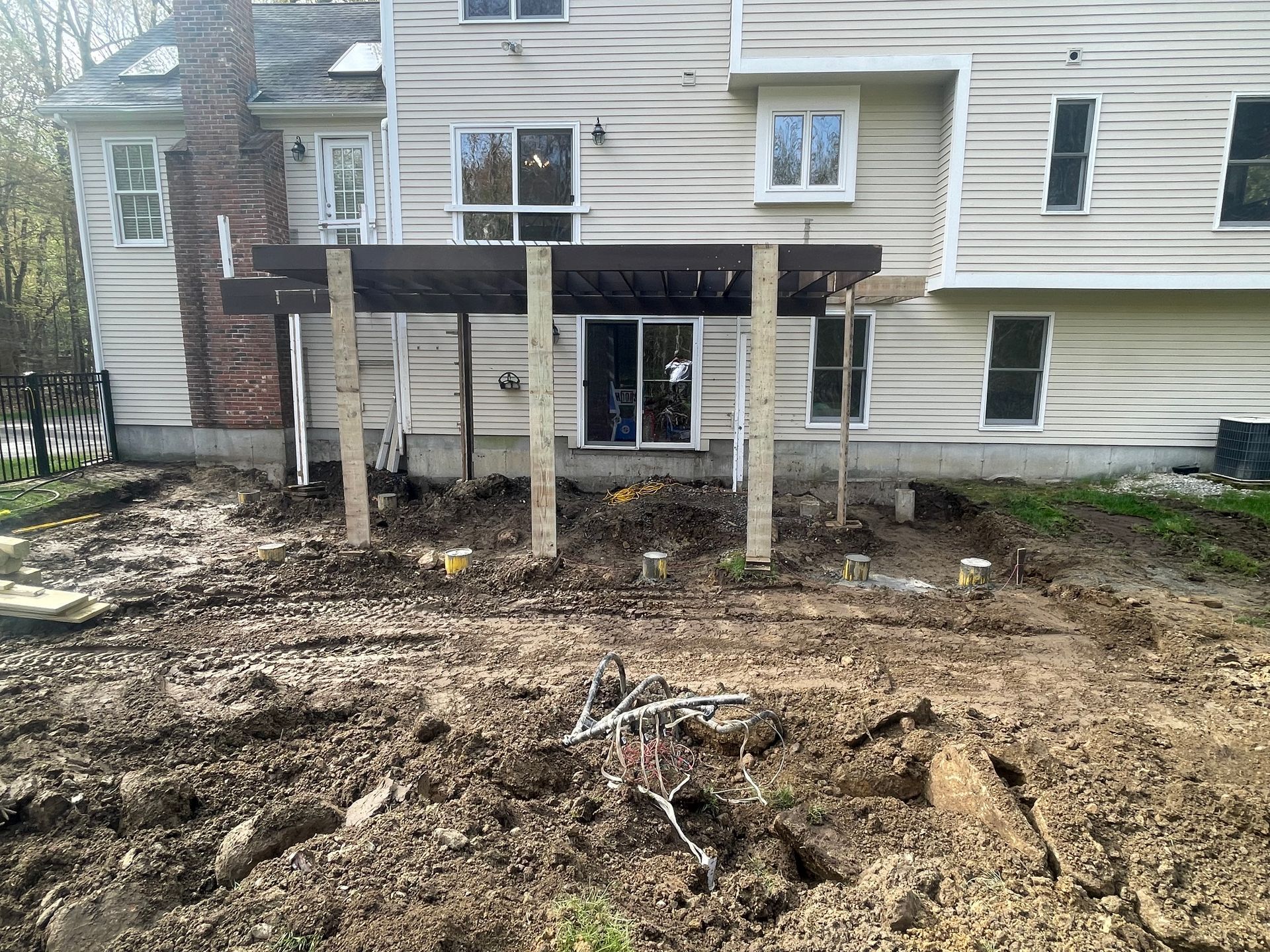 Raised wooden deck under construction behind a beige house, with dirt and support posts in front.