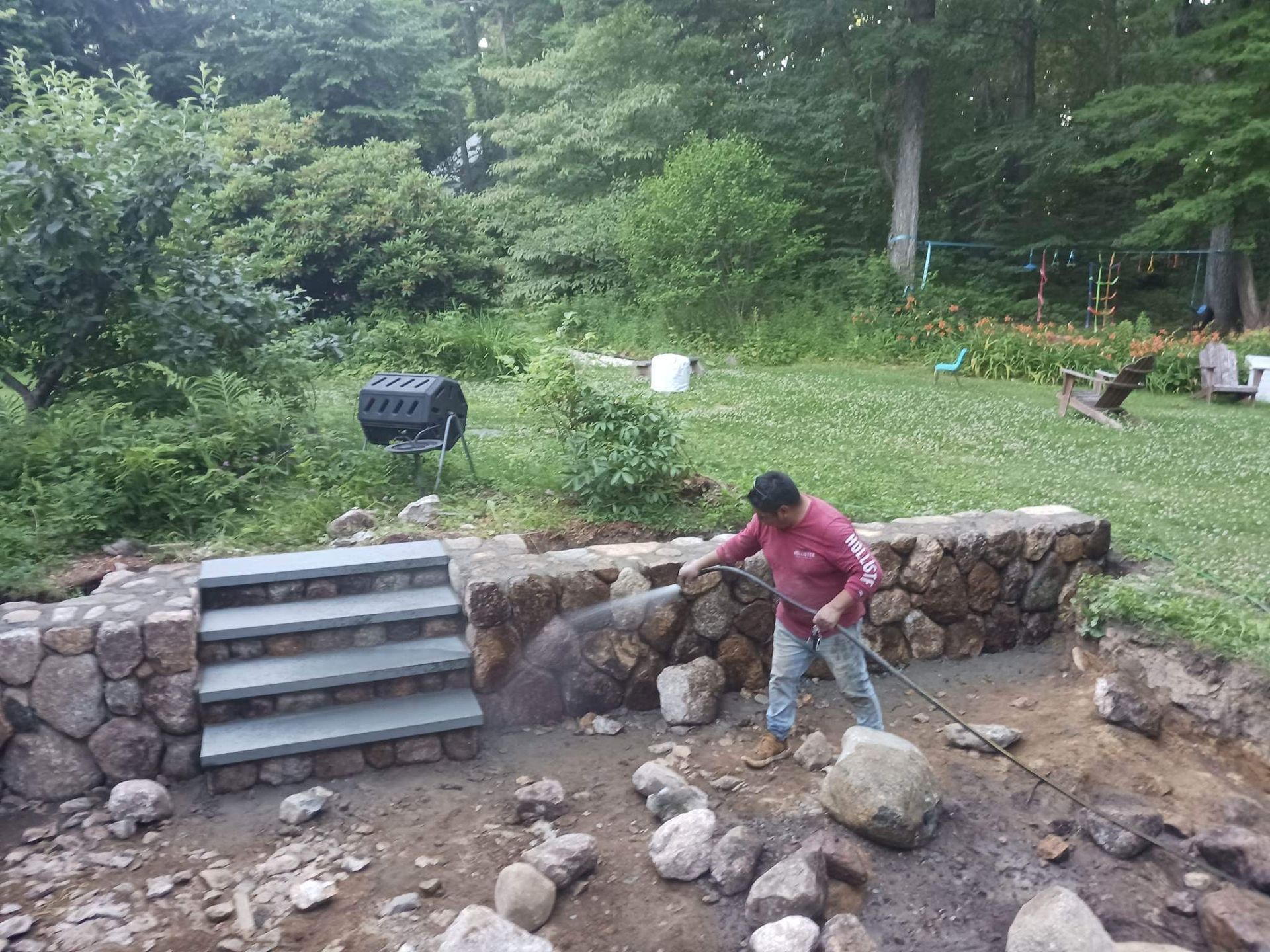 Person in a red shirt moving rocks beside a stone retaining wall in a wooded yard.