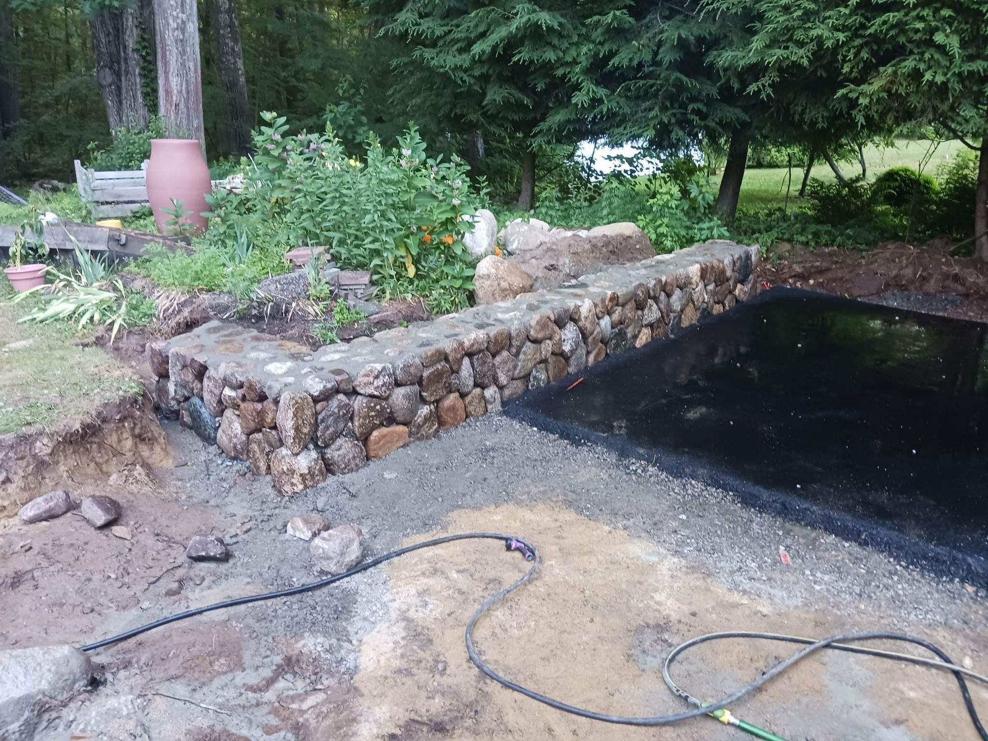 Stone-edged garden pond with plants and a hose on a gravel patio, beside a dark water feature