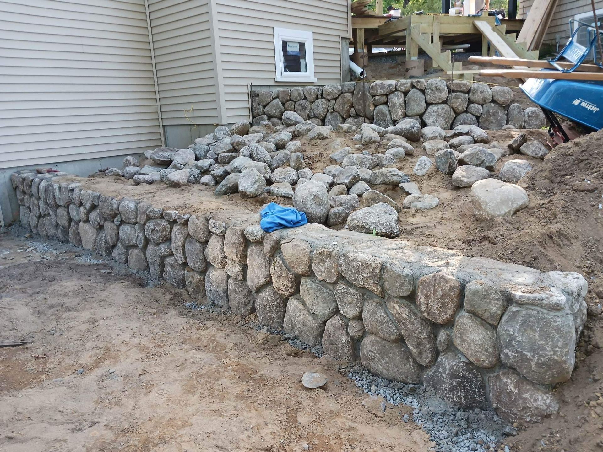 Stone retaining wall and steps under construction beside a house, with a blue tarp on the rocks.