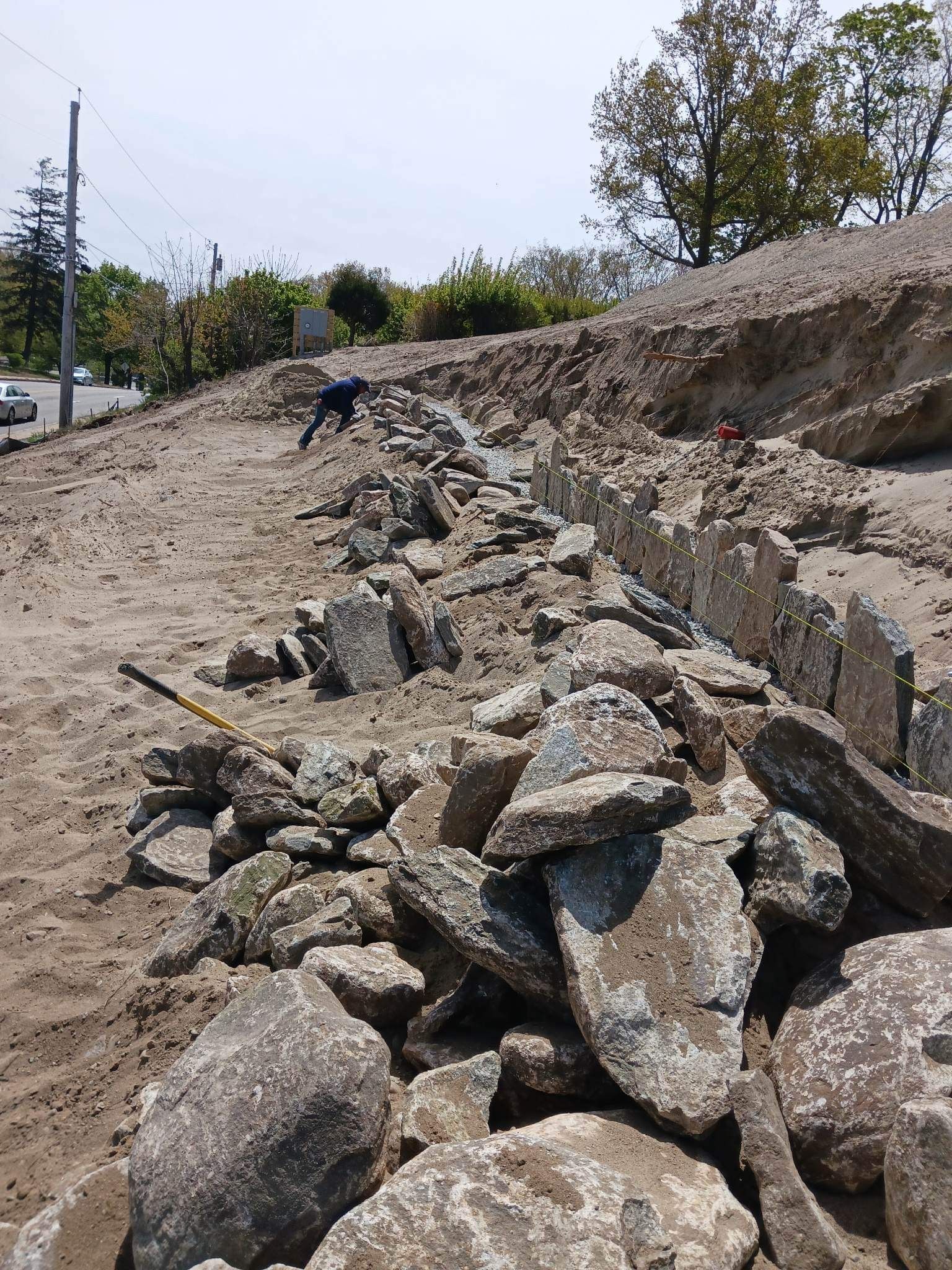 Large pile of broken concrete and rocks lining a dirt roadside embankment near parked cars and trees