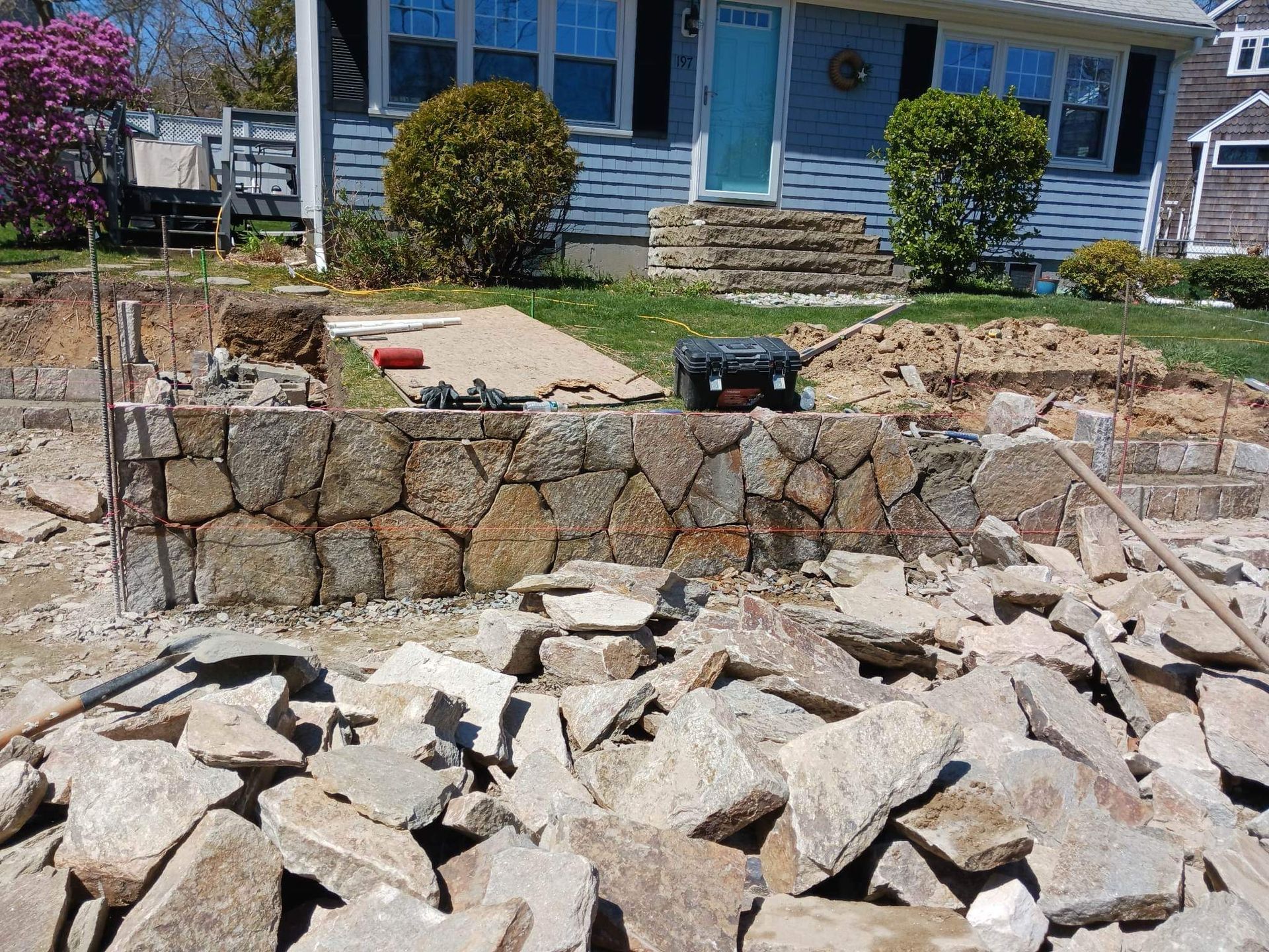 Stone retaining wall under construction in front of a blue house, with rubble in the foreground.