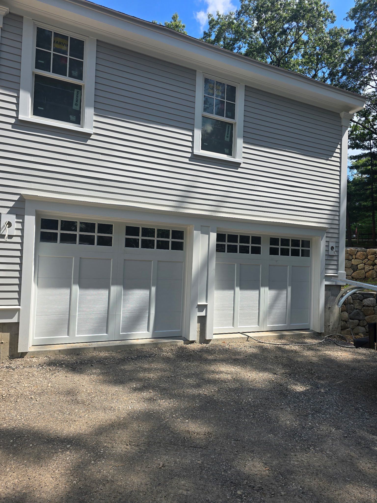 Two-car garage with white doors, gray siding, and gravel driveway. Windows above the garage.
