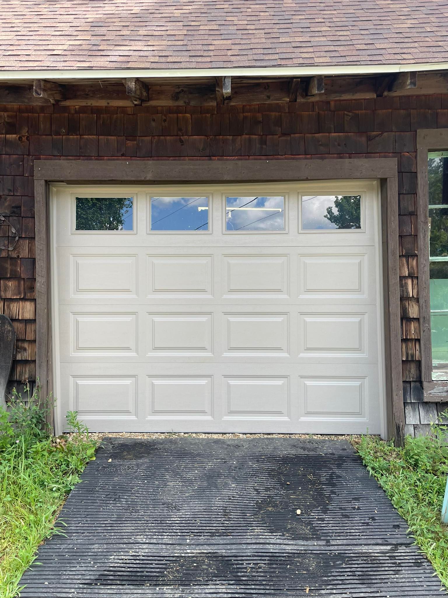 Tan garage door with windows, framed by wood siding, and leading onto a textured black walkway.