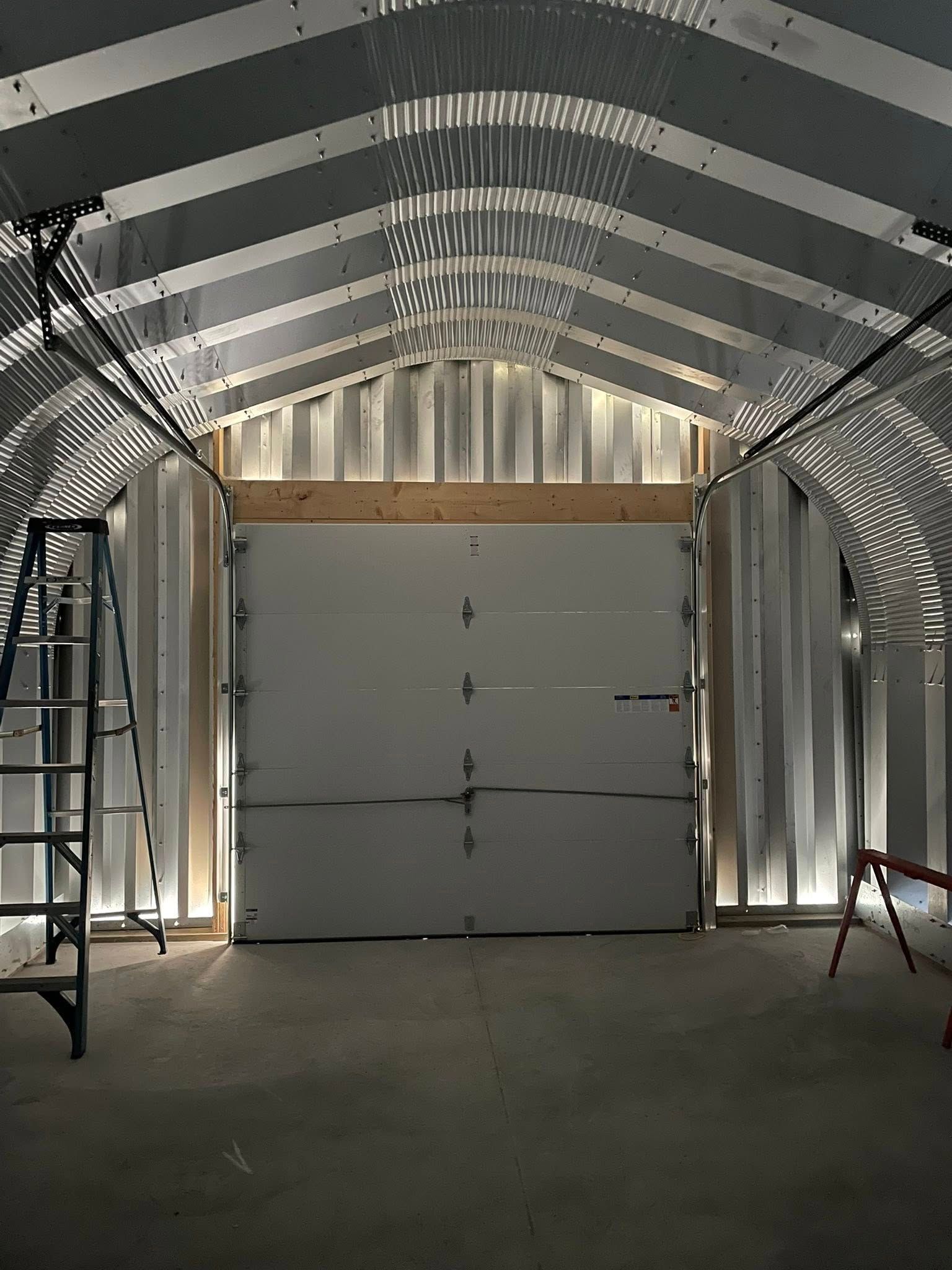 Interior view of a metal building under construction with a garage door framed by wooden beams.