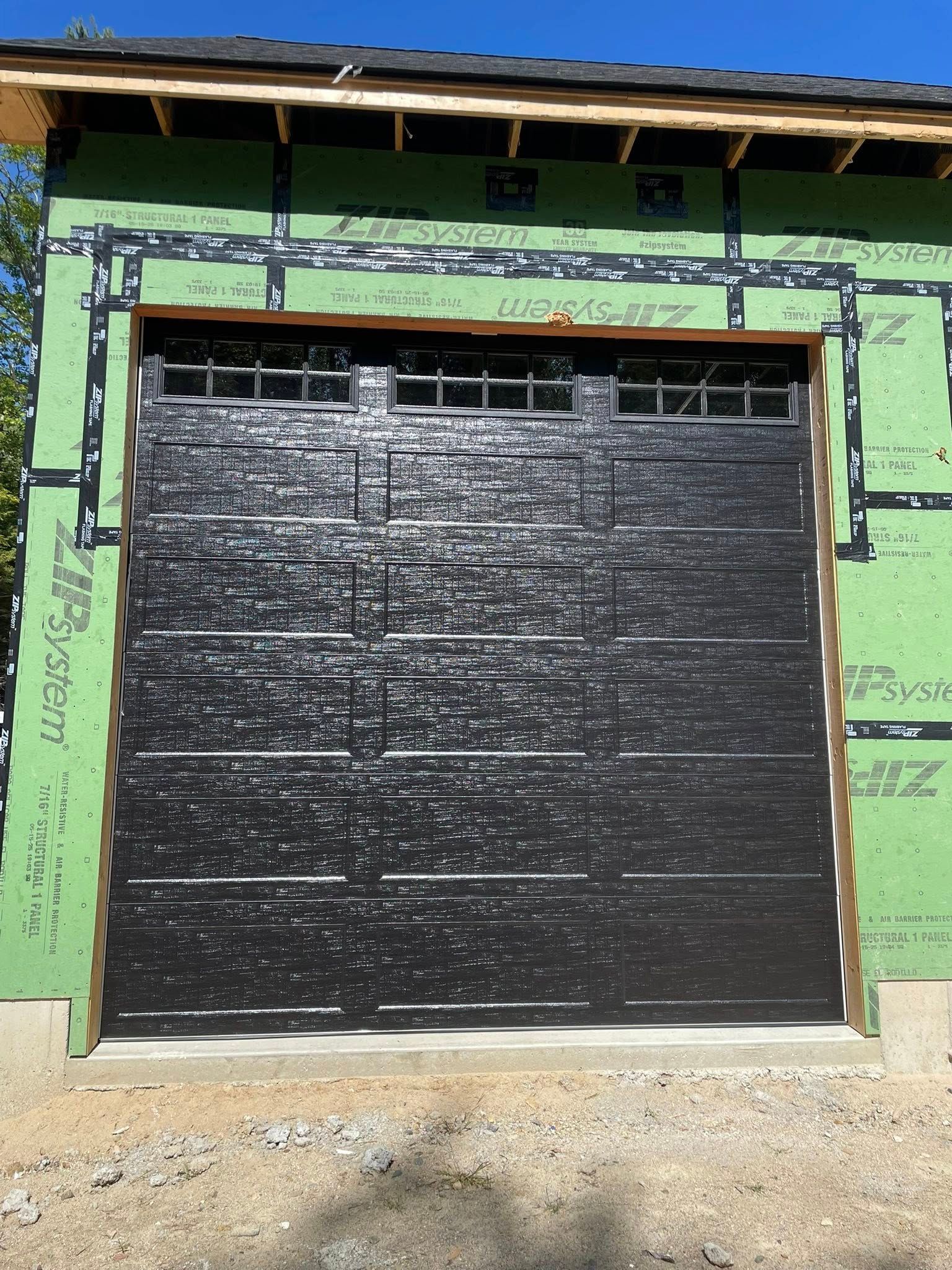 Black garage door on a new building, surrounded by green insulation and brown trim.