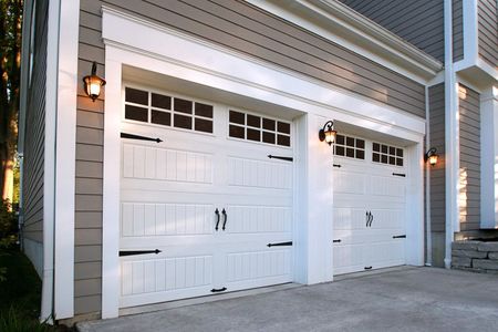 White garage doors with black hardware and windows, flanked by sconces. Gray siding.