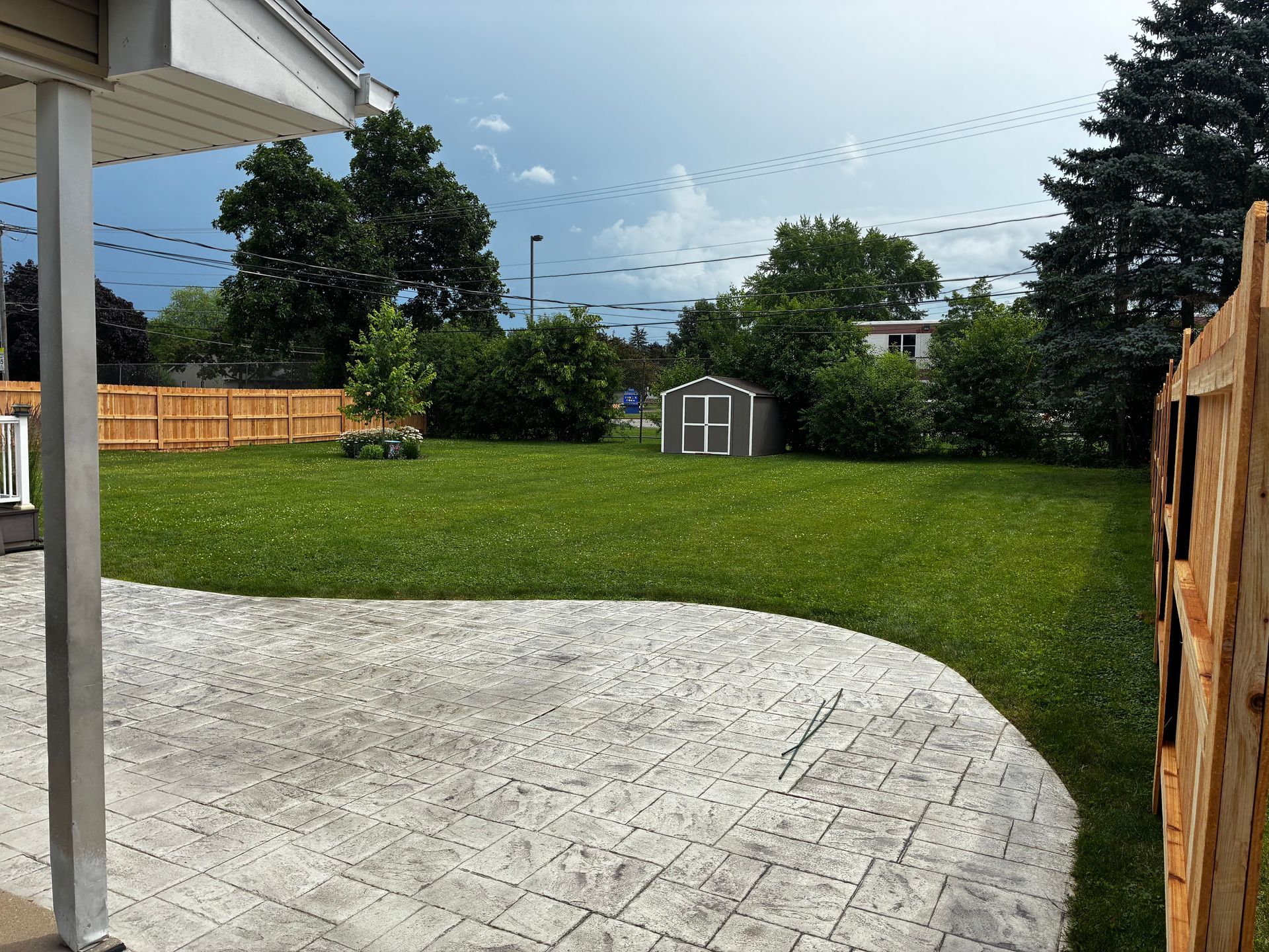 Backyard with a green lawn, concrete patio, wooden fence, and small shed under a partly cloudy sky